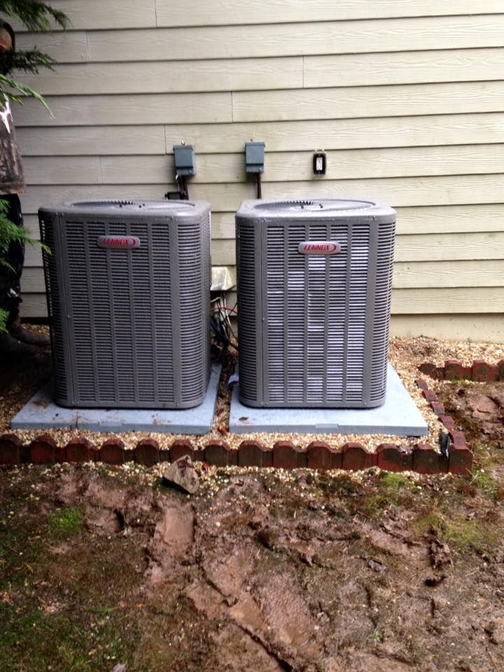 Two gray outdoor HVAC units on concrete pads beside a house wall, with dirt and leaves in the foreground
