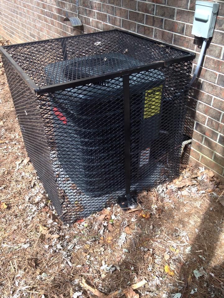 Black outdoor air conditioner unit inside a wire mesh cage beside a brick wall