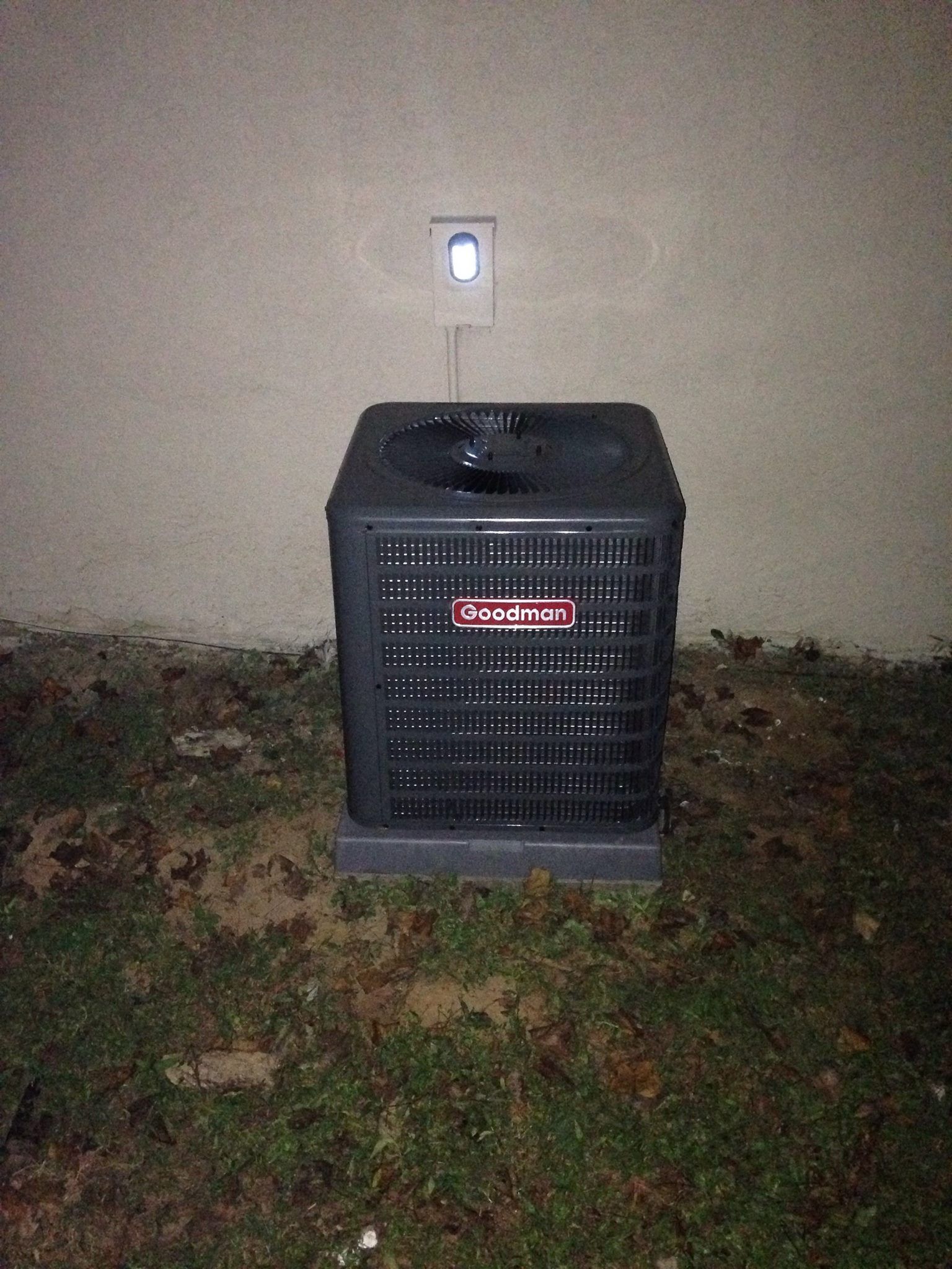 Black outdoor heater under a wall outlet at night, standing on grass and leaves.
