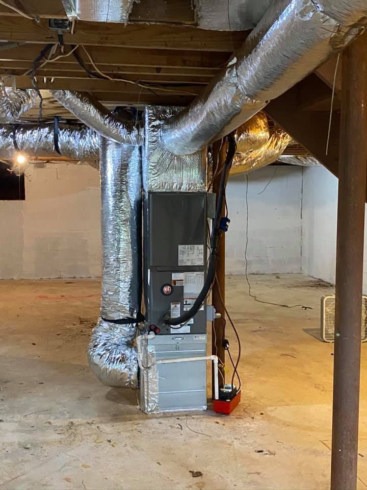 Basement HVAC furnace with silver ductwork and exposed beams in an unfinished utility space