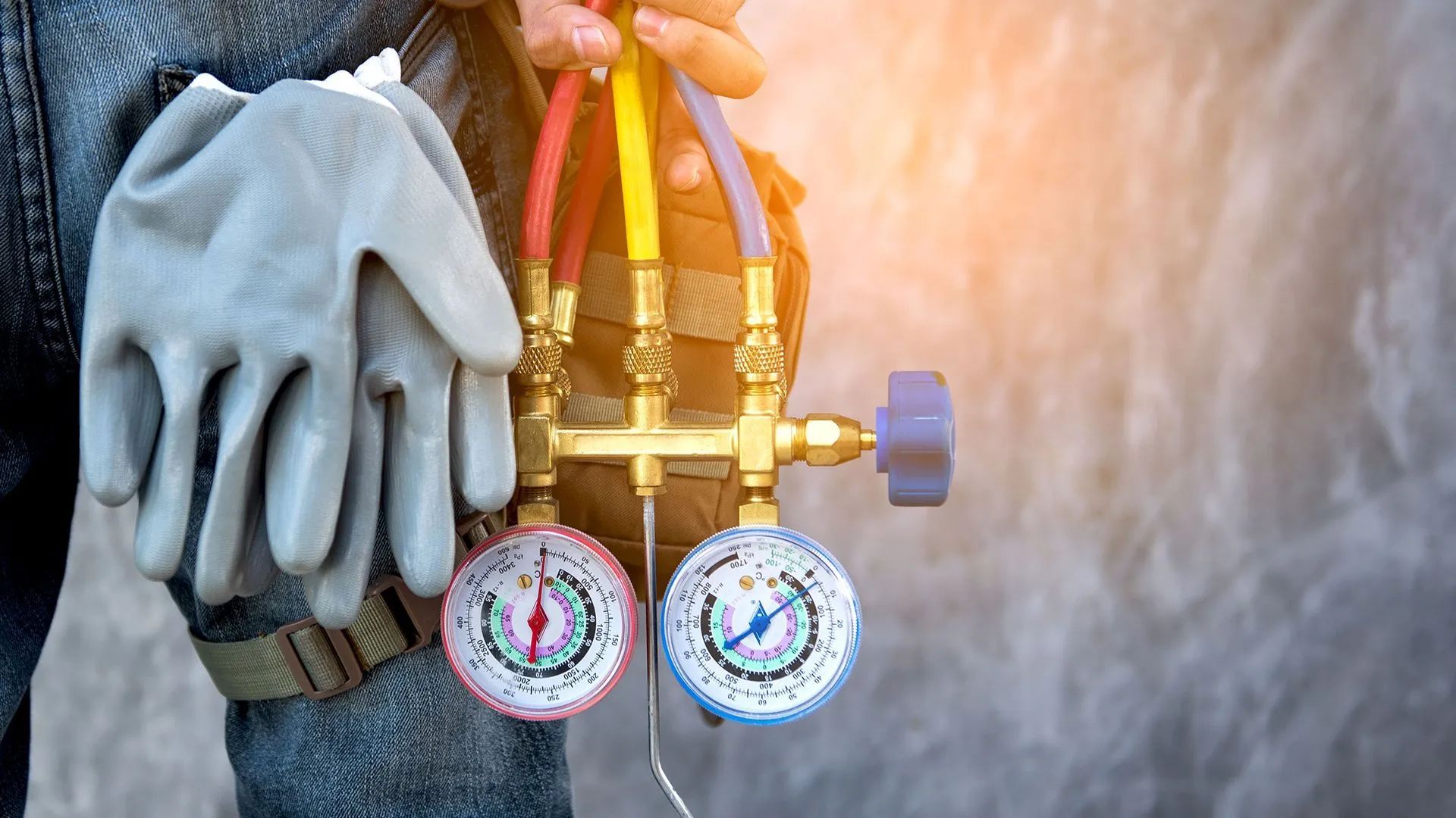 Gloved hand holding HVAC gauges and hoses against an outdoor unit