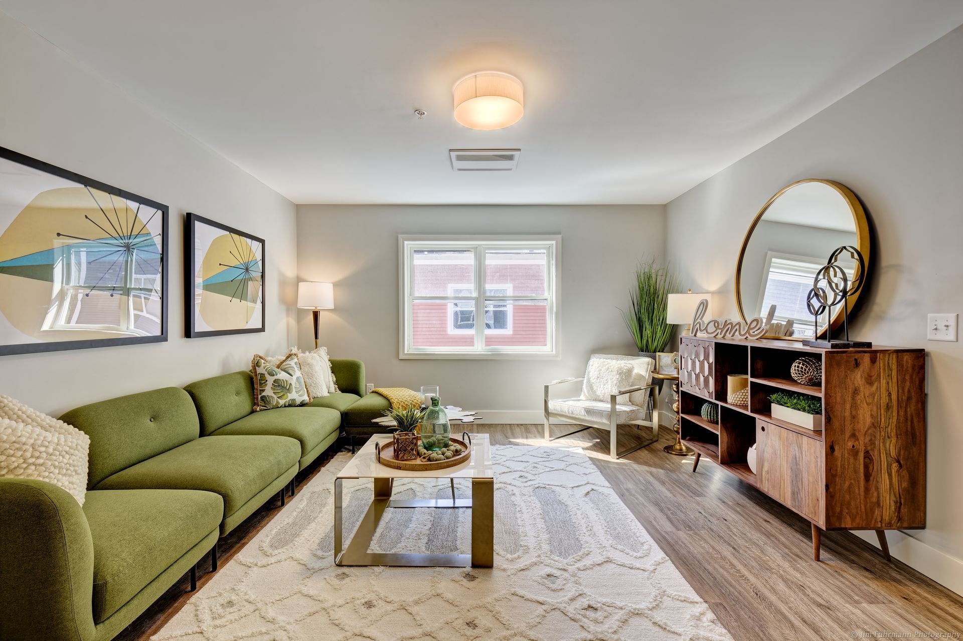 A living room with a green sectional sofa, wooden cabinet, and a large round mirror.
