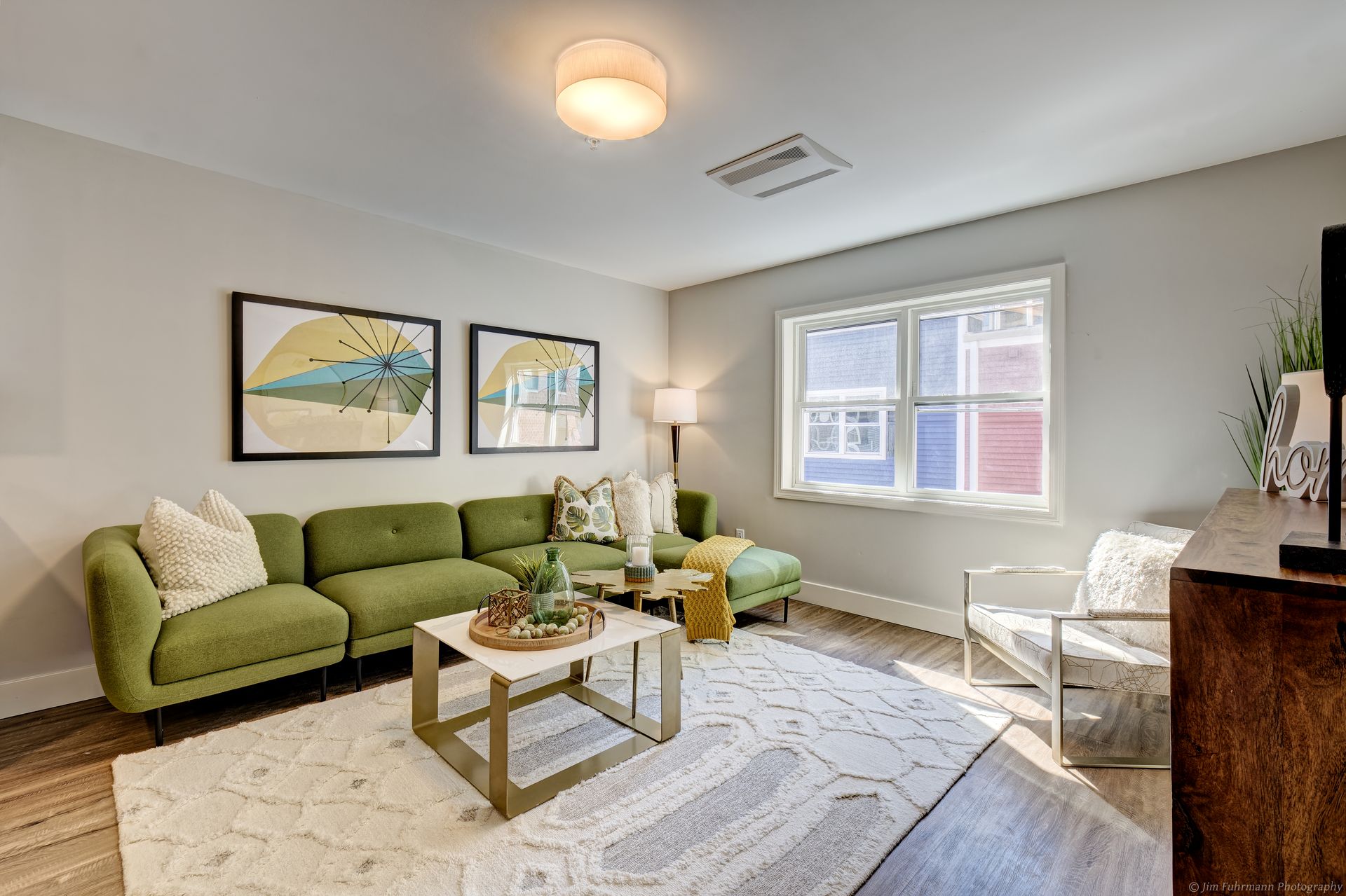 A modern living room with a green sectional sofa, white rug, and artwork. Sunlight streams through a window onto the wood floor.
