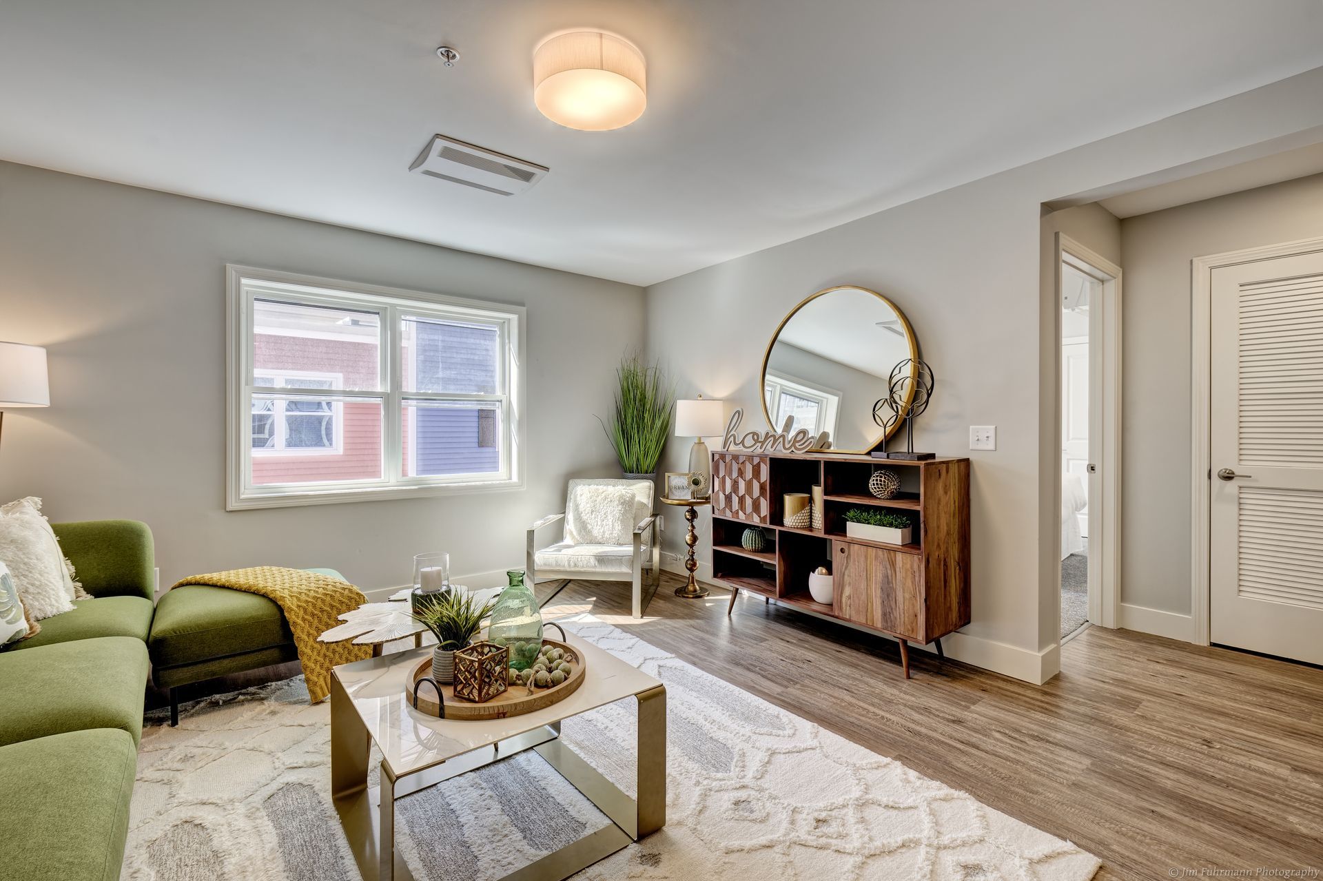 Living room with green couch, wooden cabinet, round mirror, and window. Neutral walls, wooden floors, and a rug.