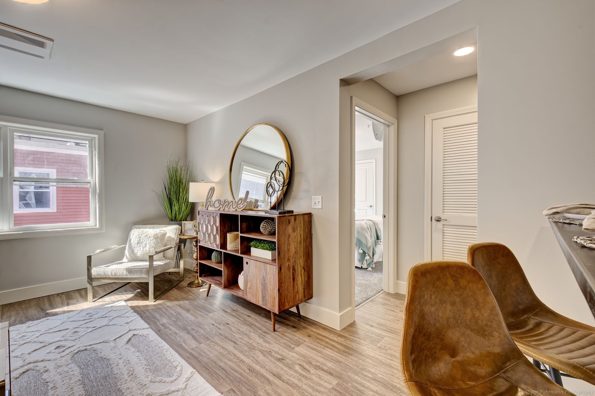 A living room with a mid-century modern aesthetic featuring a wooden cabinet, a mirror, and a view into a bedroom.
