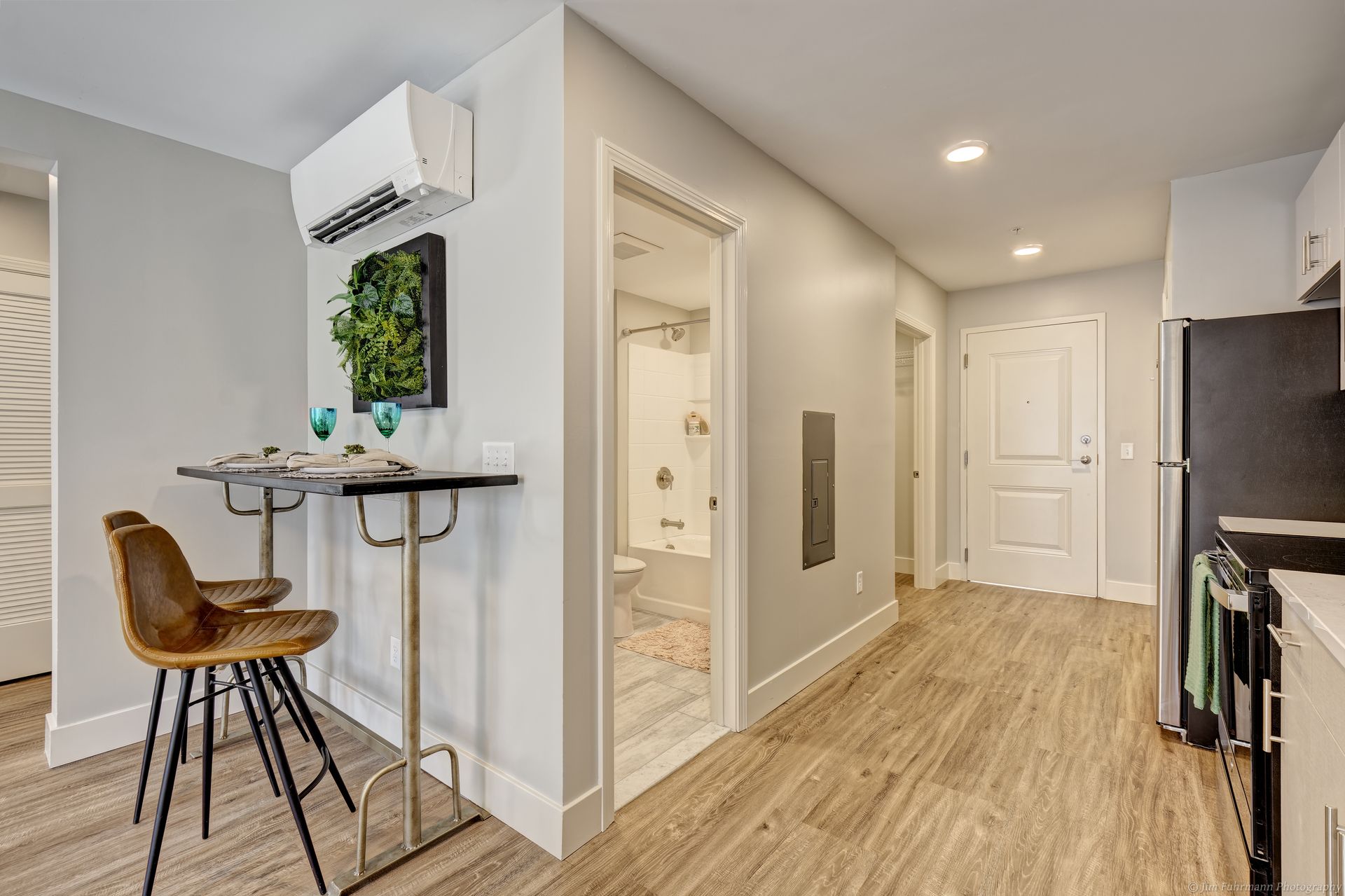 A small apartment interior with a dining table, kitchen, hallway, and open doorway to a bathroom. Light walls and wood-look flooring.