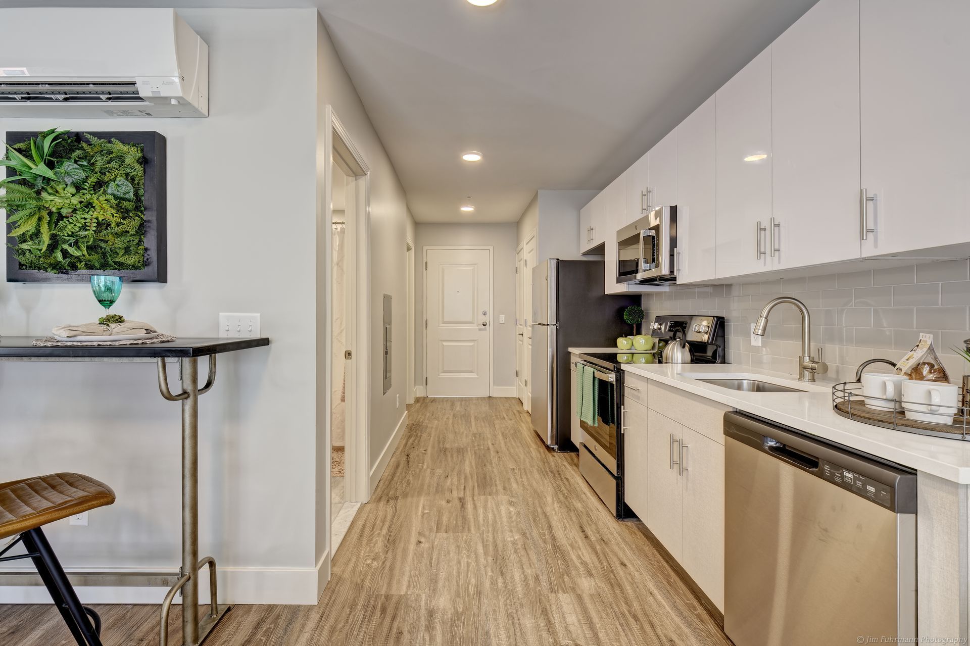 Narrow modern apartment kitchen with white cabinets, stainless steel appliances, and wood-look flooring. A small table with a stool is in the foreground.