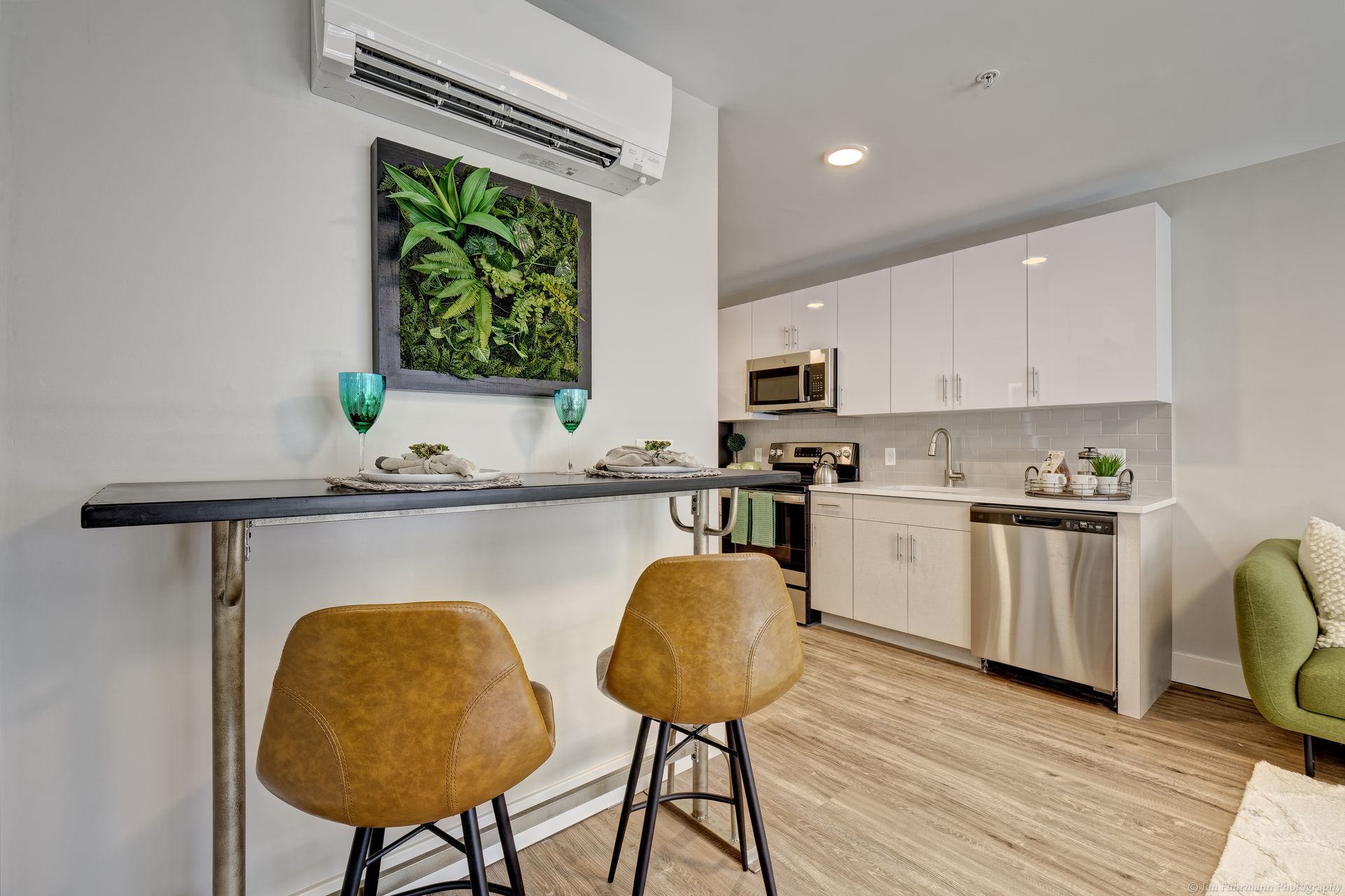 A modern kitchen with a breakfast bar and two brown leather stools. White cabinets, stainless steel appliances, and a green plant wall art piece are featured.
