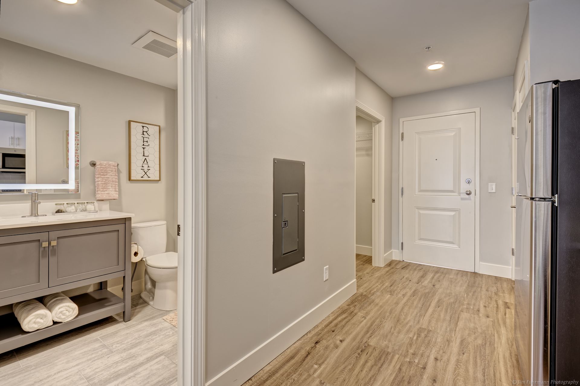 Hallway with a bathroom on the left, a door straight ahead, and a refrigerator on the right. Light gray walls, wood-look floors.