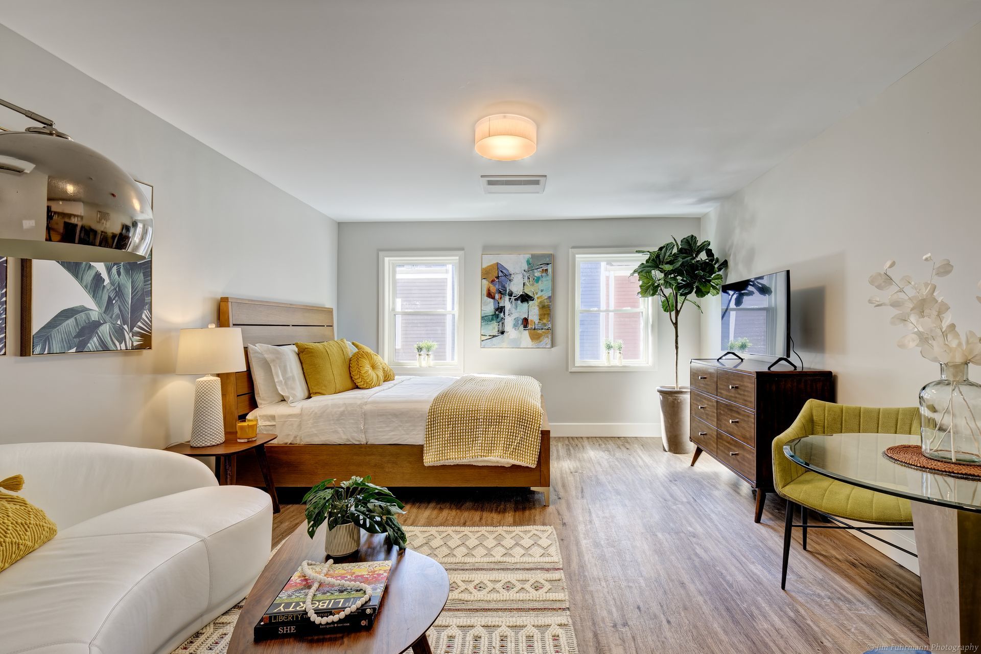 Cozy studio apartment with bed, sofa, TV, and dining table. Light gray walls, wood flooring, and natural light.