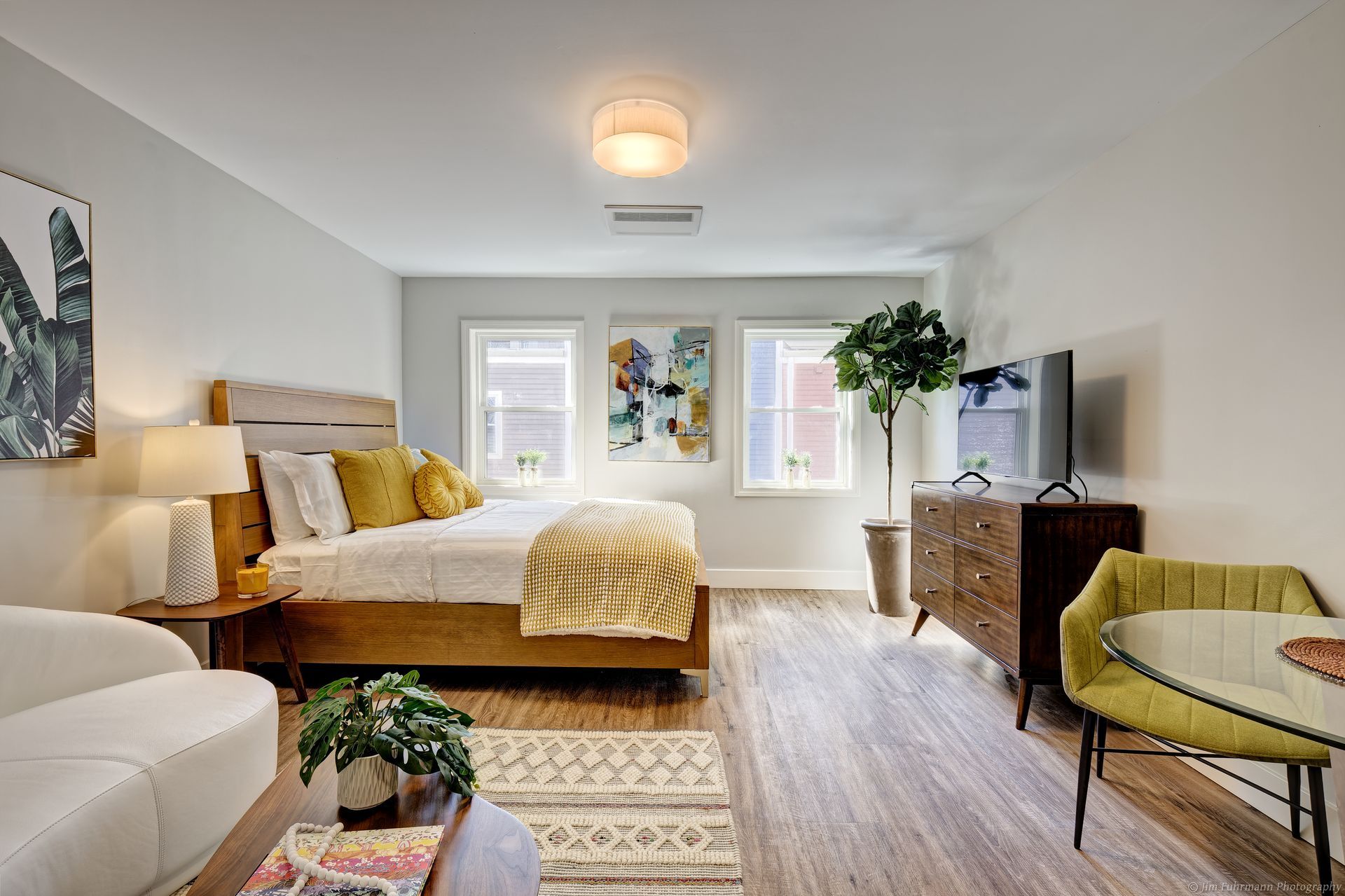 A modern studio apartment with a bed, TV, small dining area, and couch, decorated with neutral tones and pops of yellow.