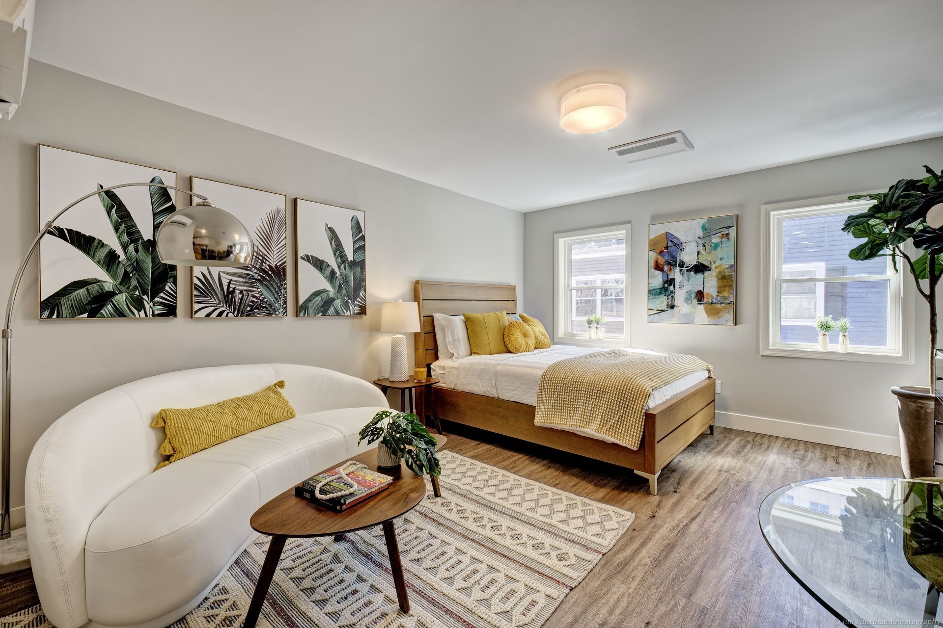 A bright, modern studio apartment. Features a bed, sofa, coffee table, art, and large windows. White, gold, and green colors throughout.