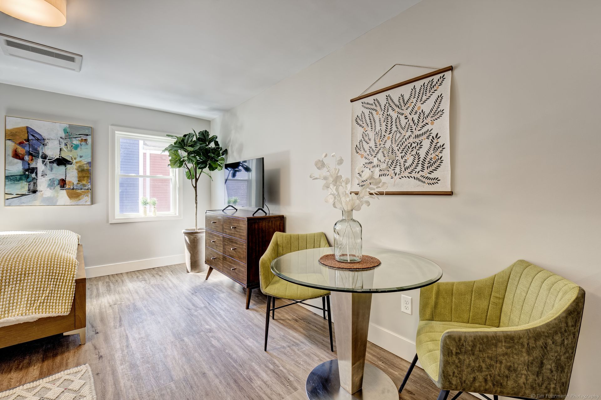 A small studio apartment with hardwood floors. Features a bed, table with two chairs, a dresser, and artwork. Light green and gold accents.