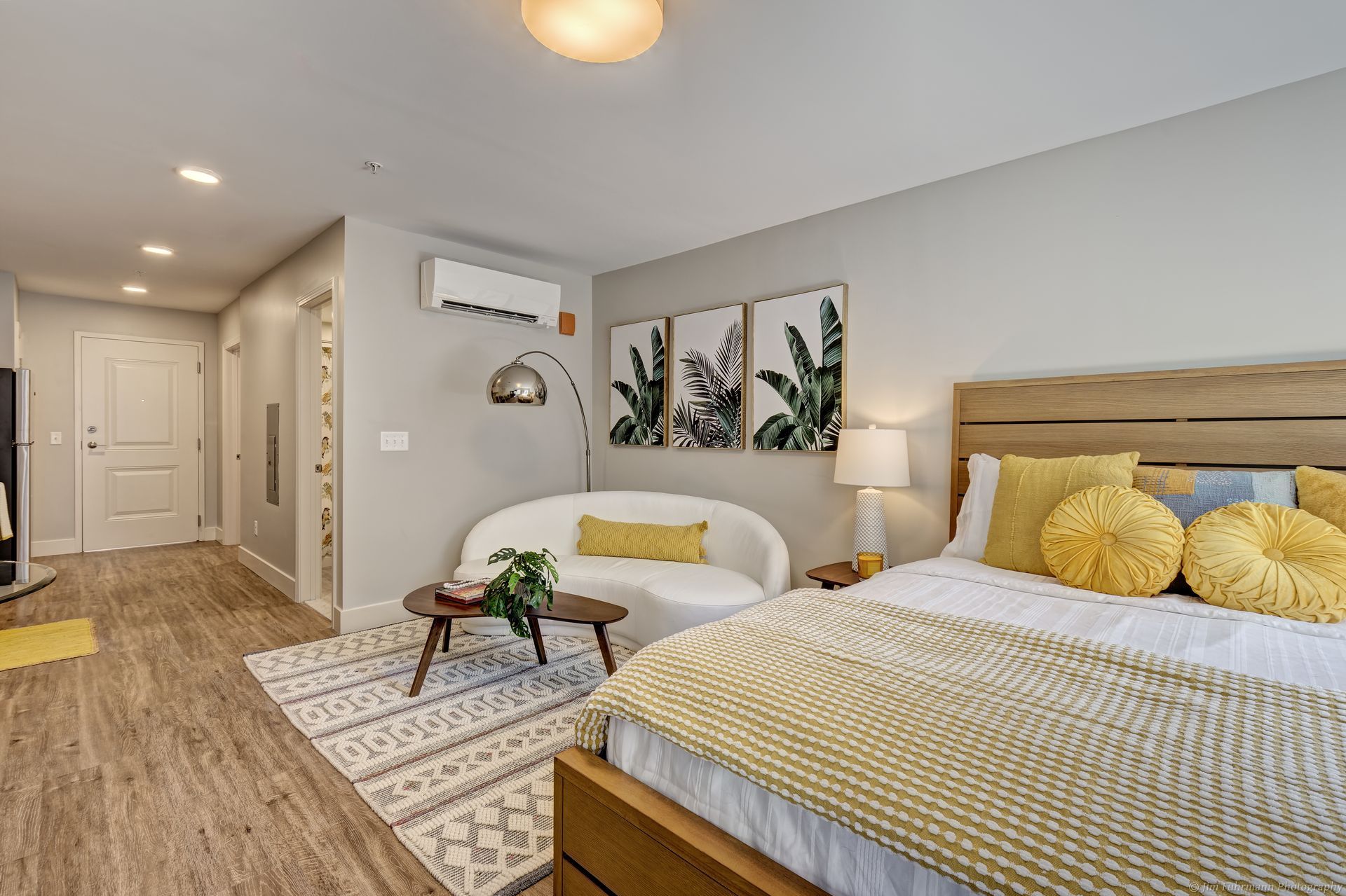 A bright, modern studio apartment with a bed, small sofa, and artwork. Yellow and white accents are used throughout the space.