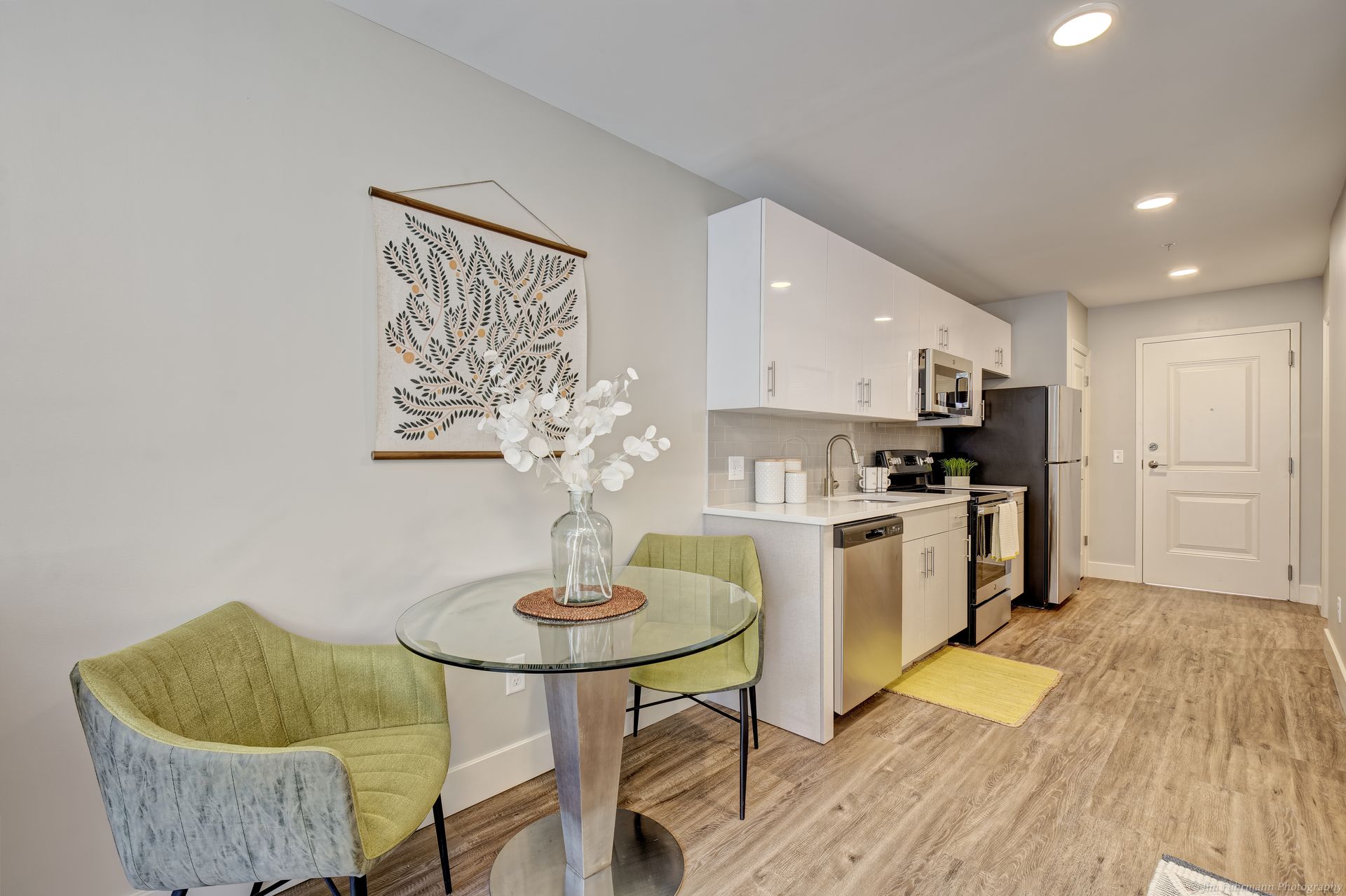 Small kitchen with a round table and two green chairs. White cabinets, stainless steel appliances, and a door are visible.