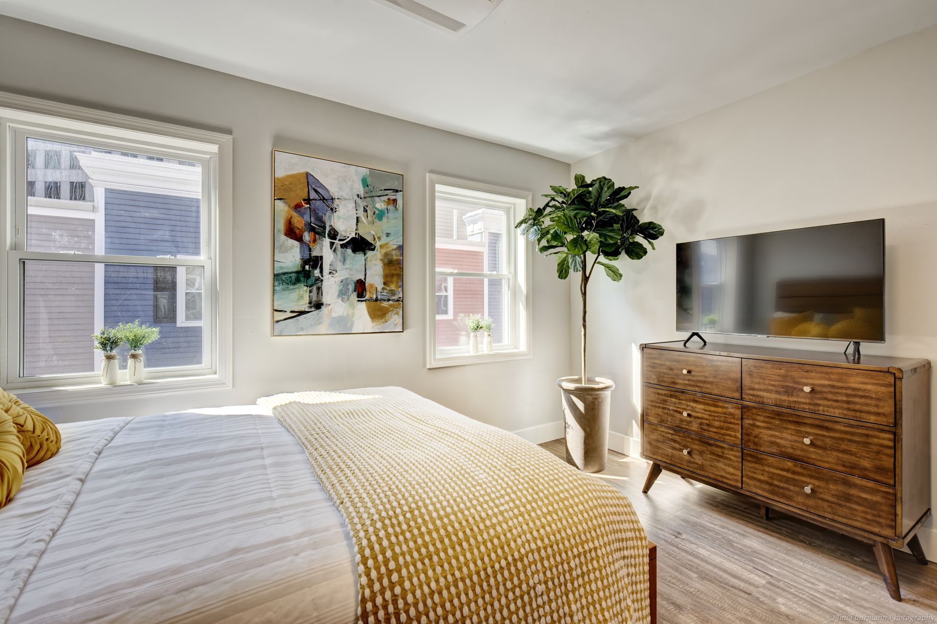 Bedroom with a bed, dresser with a TV, and a tall potted plant. Two windows with light streaming in.
