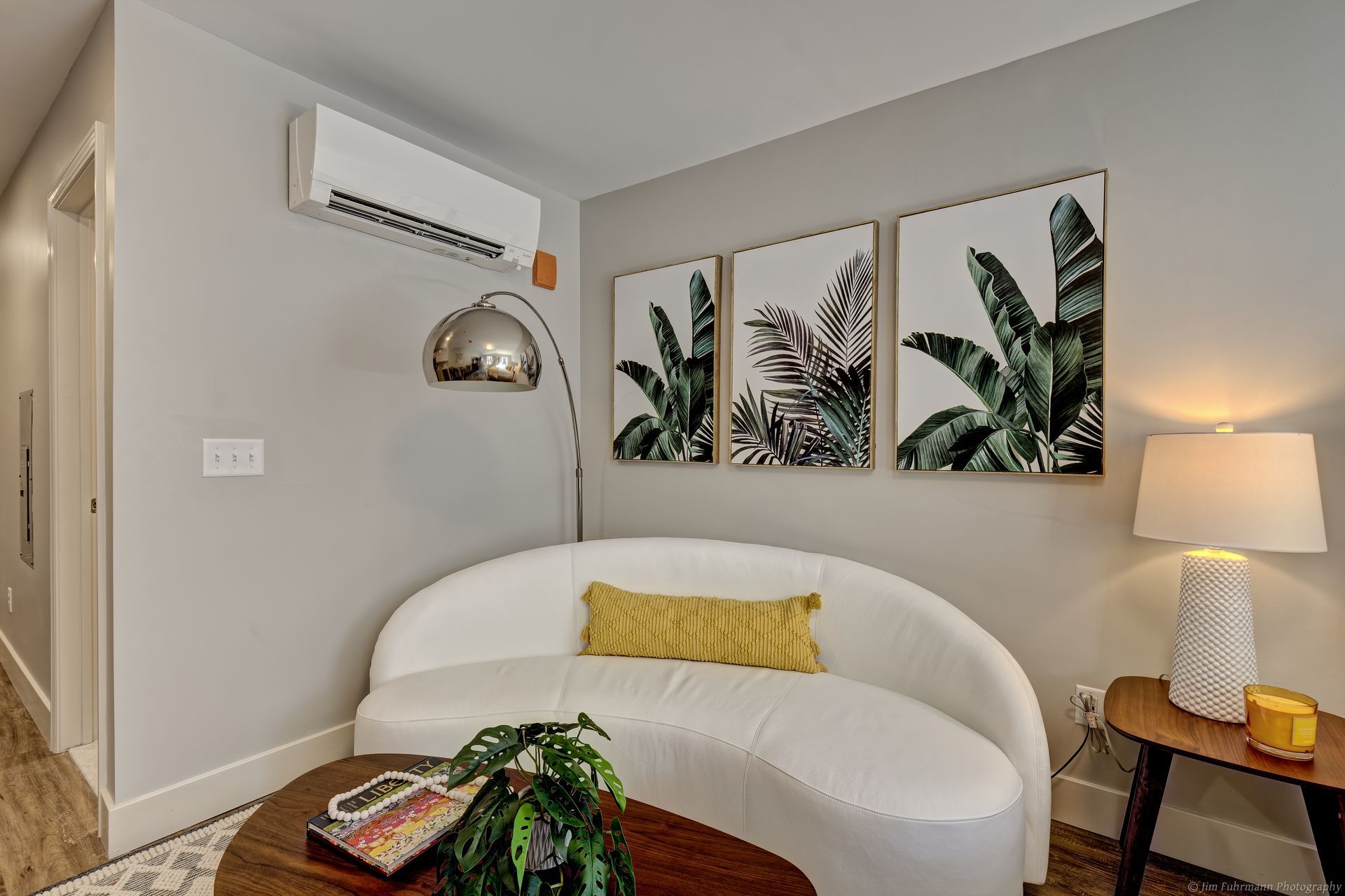 A cozy living space with a white sofa, tropical artwork, and a modern floor lamp. A side table with a lamp sits on the right.