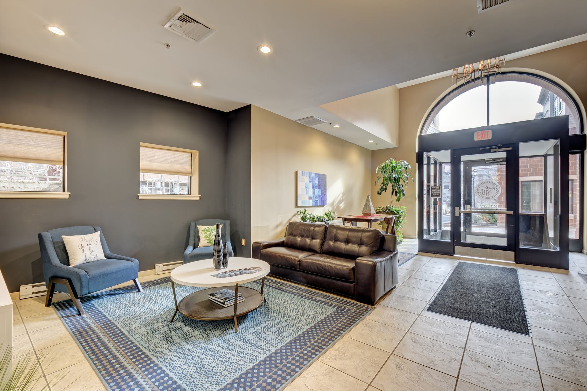 Lobby with blue chairs, a brown couch, and a marble coffee table on a patterned rug. Large arched doorway.