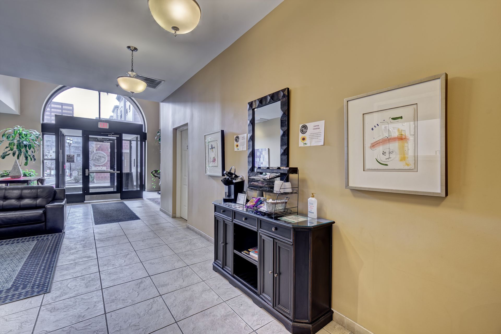 An entryway with black cabinet, mirror, and coffee station.  Doors open to the outside, a black couch to the left. Beige and white color scheme.