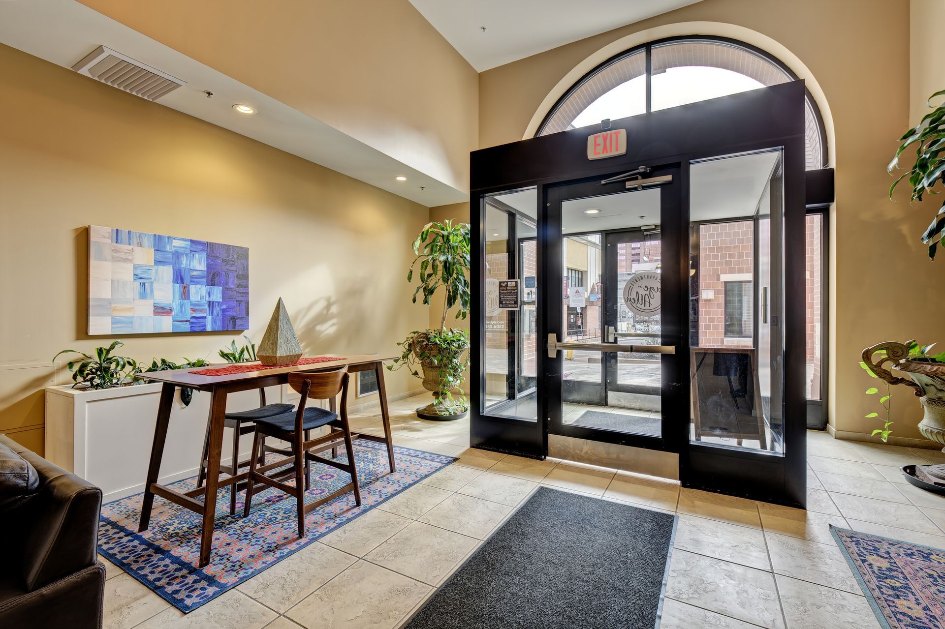 Lobby entrance with glass doors, a small table, and a painting. The room has light-colored walls and flooring, with plants.