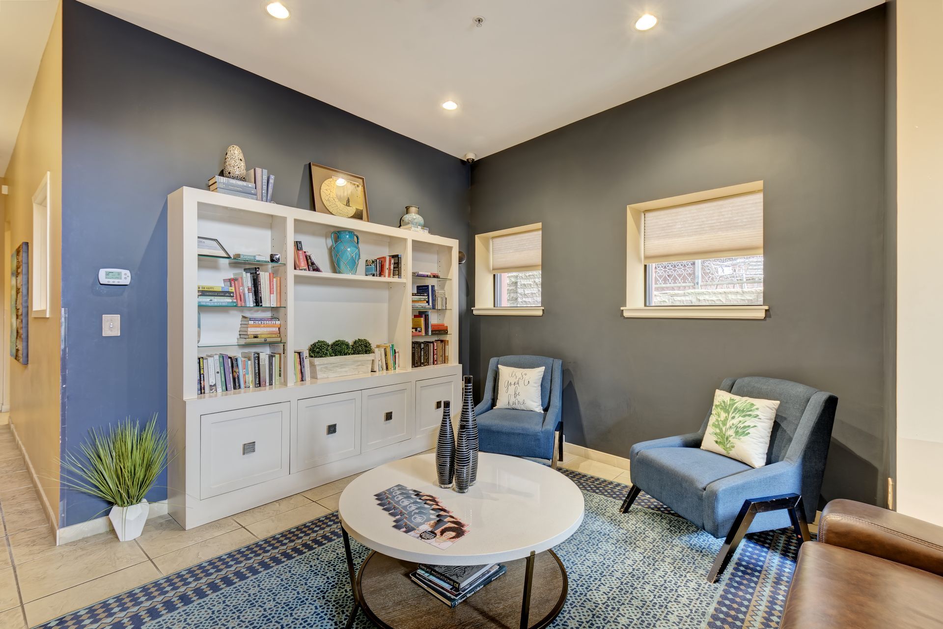 A cozy living room with blue walls, a white bookcase, and two blue armchairs. A round coffee table sits on a blue rug.