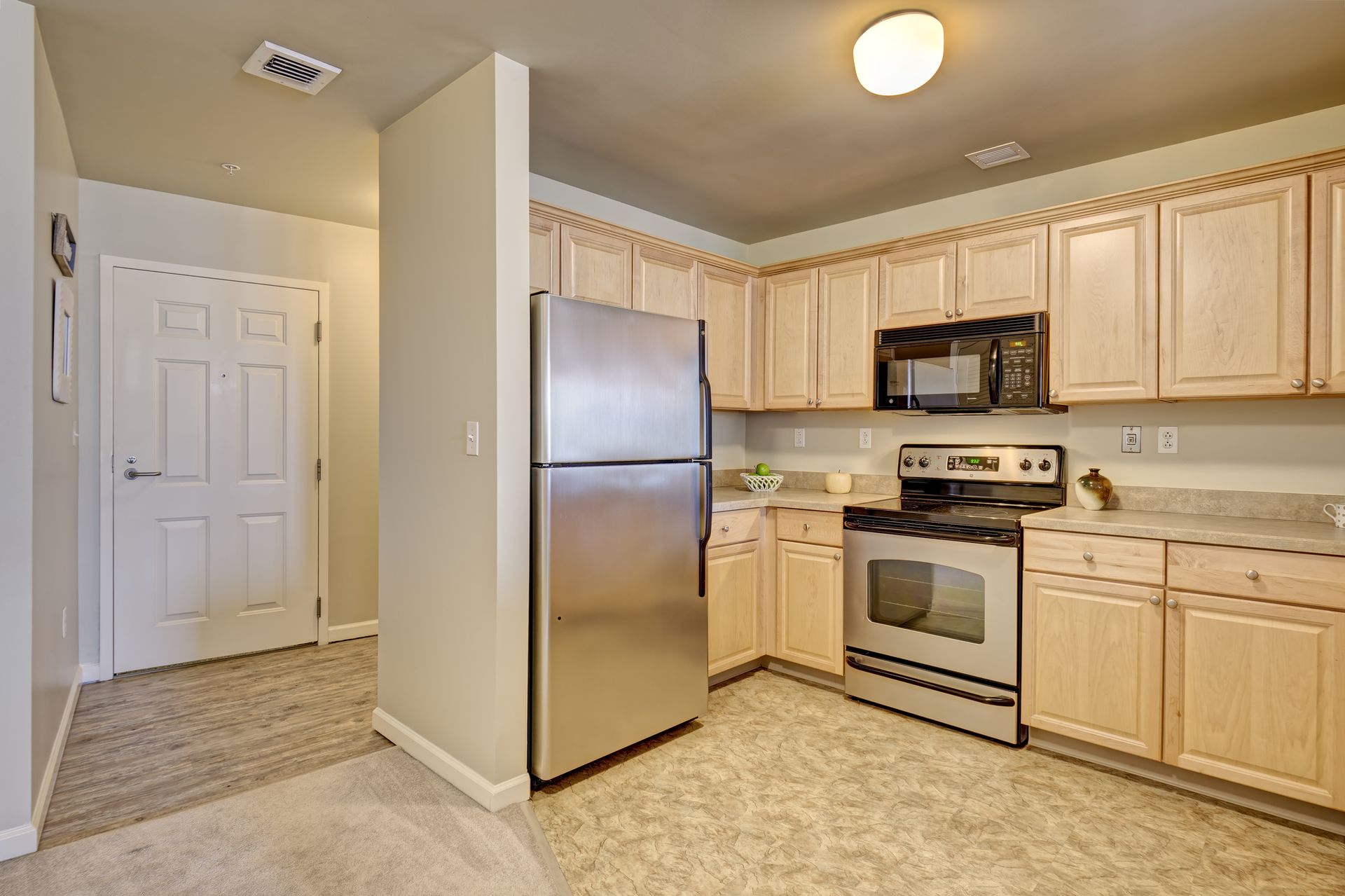 Kitchen with light wood cabinets, stainless steel appliances, and a light gray countertop. A doorway is visible on the left.