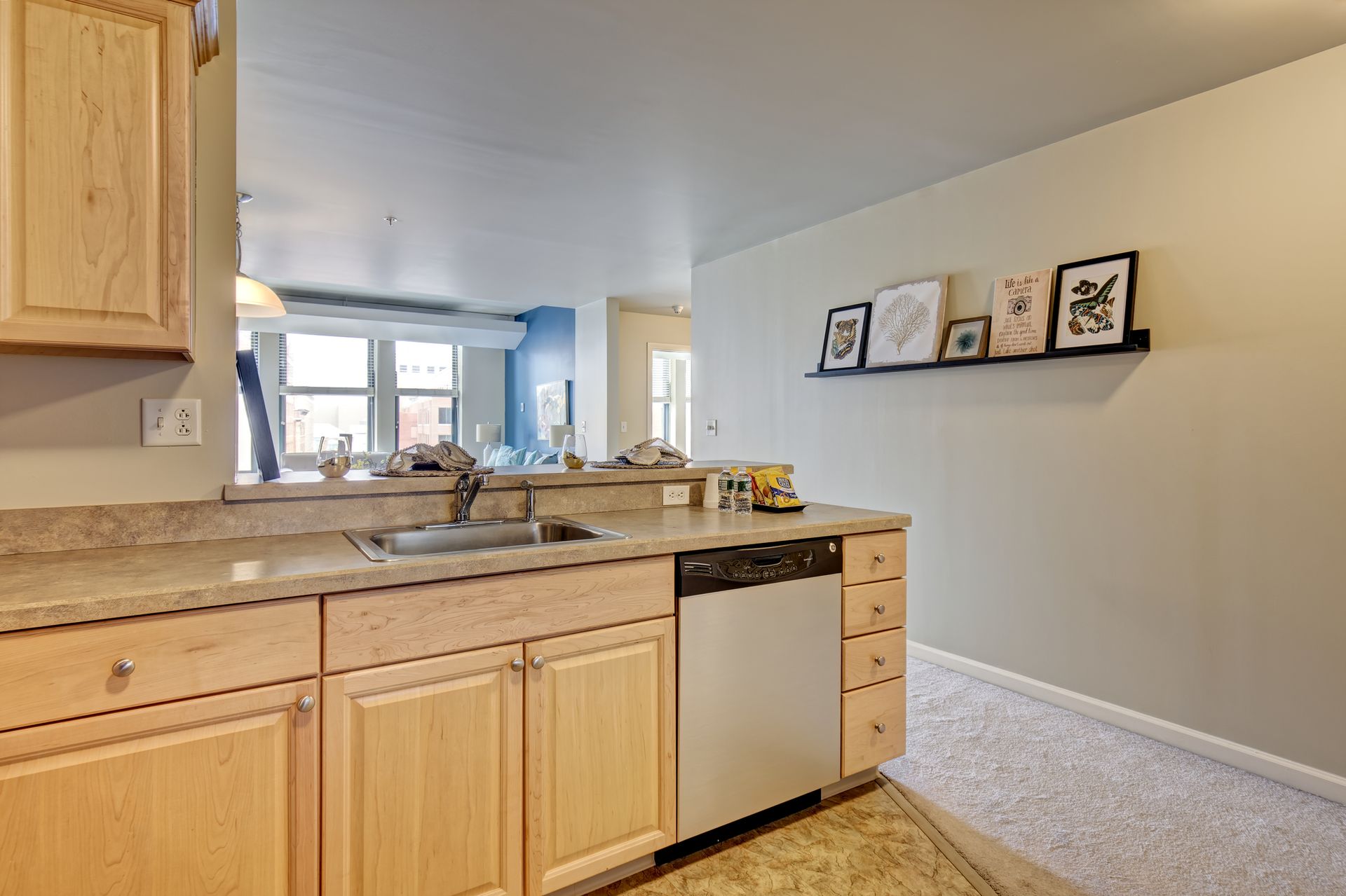 Kitchen area with light wood cabinets, a stainless steel dishwasher, and a sink. A long shelf with framed artwork is on the wall.
