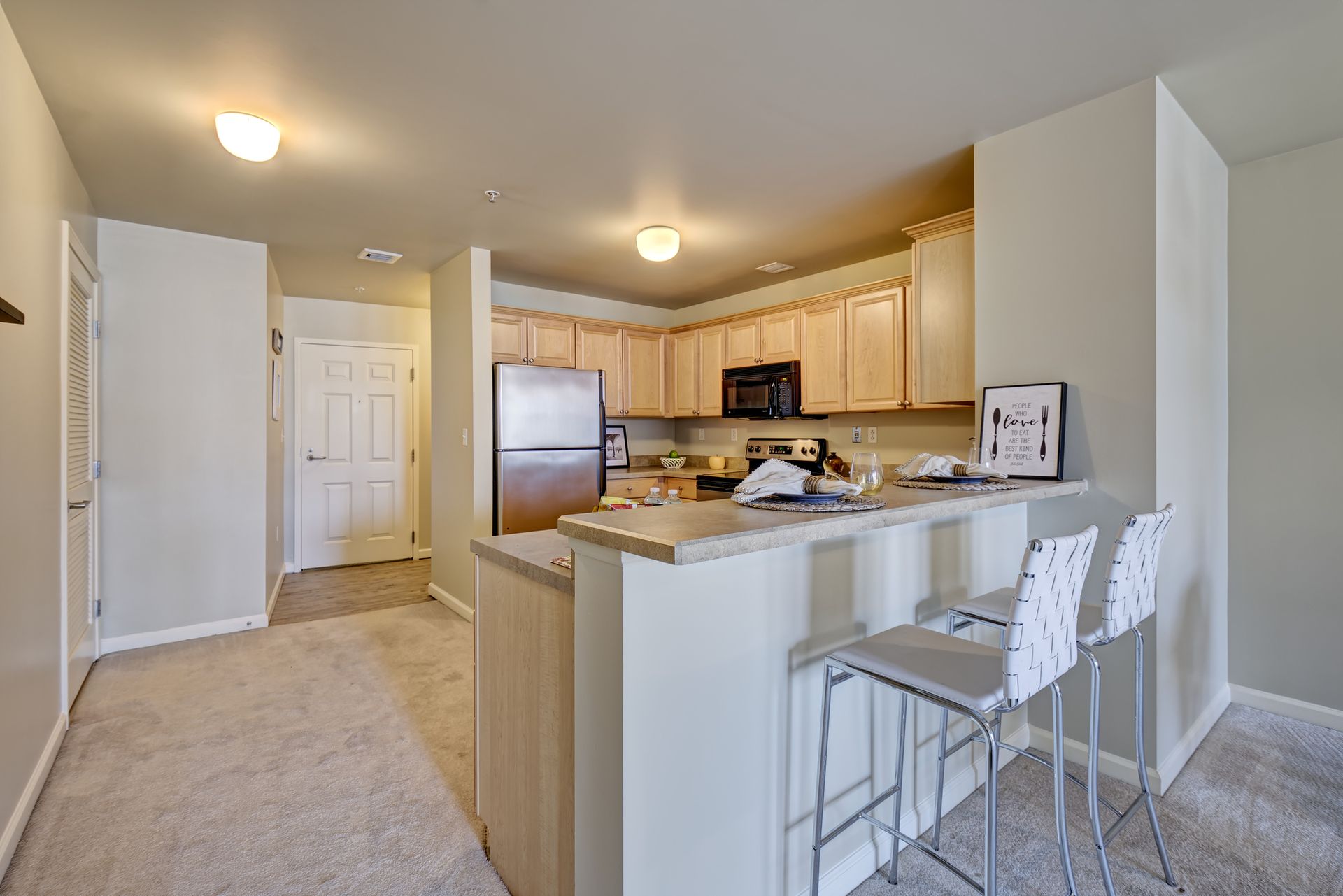Kitchen with light cabinets, stainless steel fridge, breakfast bar with stools, and carpet.