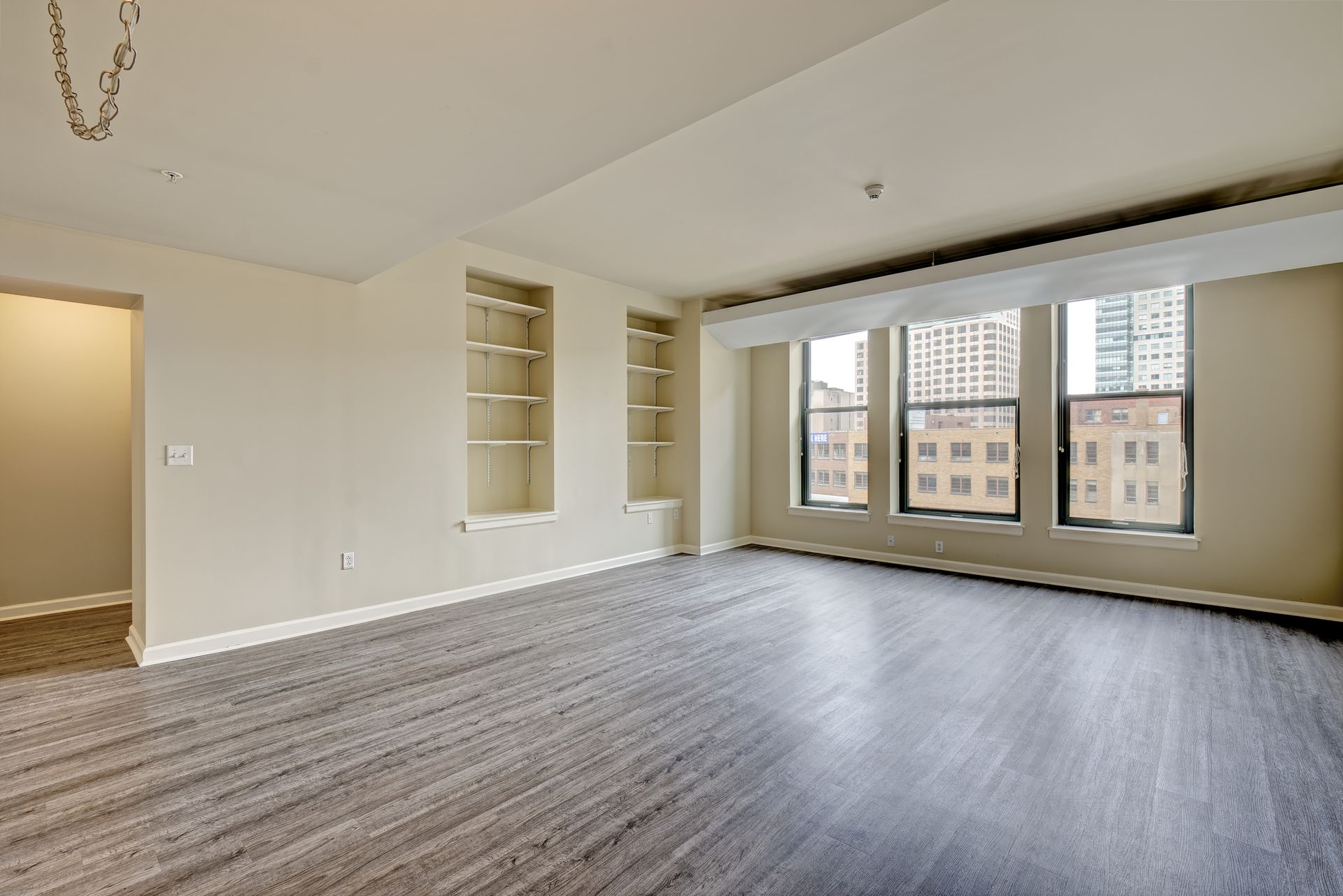 Empty living room with light walls, built-in shelves, large windows, and carpet flooring.