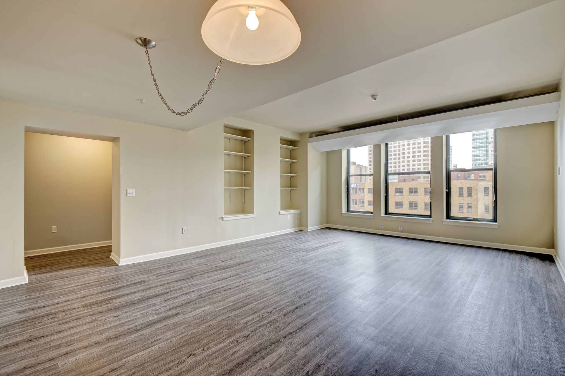 Empty living room with gray carpet, three windows, built-in shelves, and a hanging light fixture.