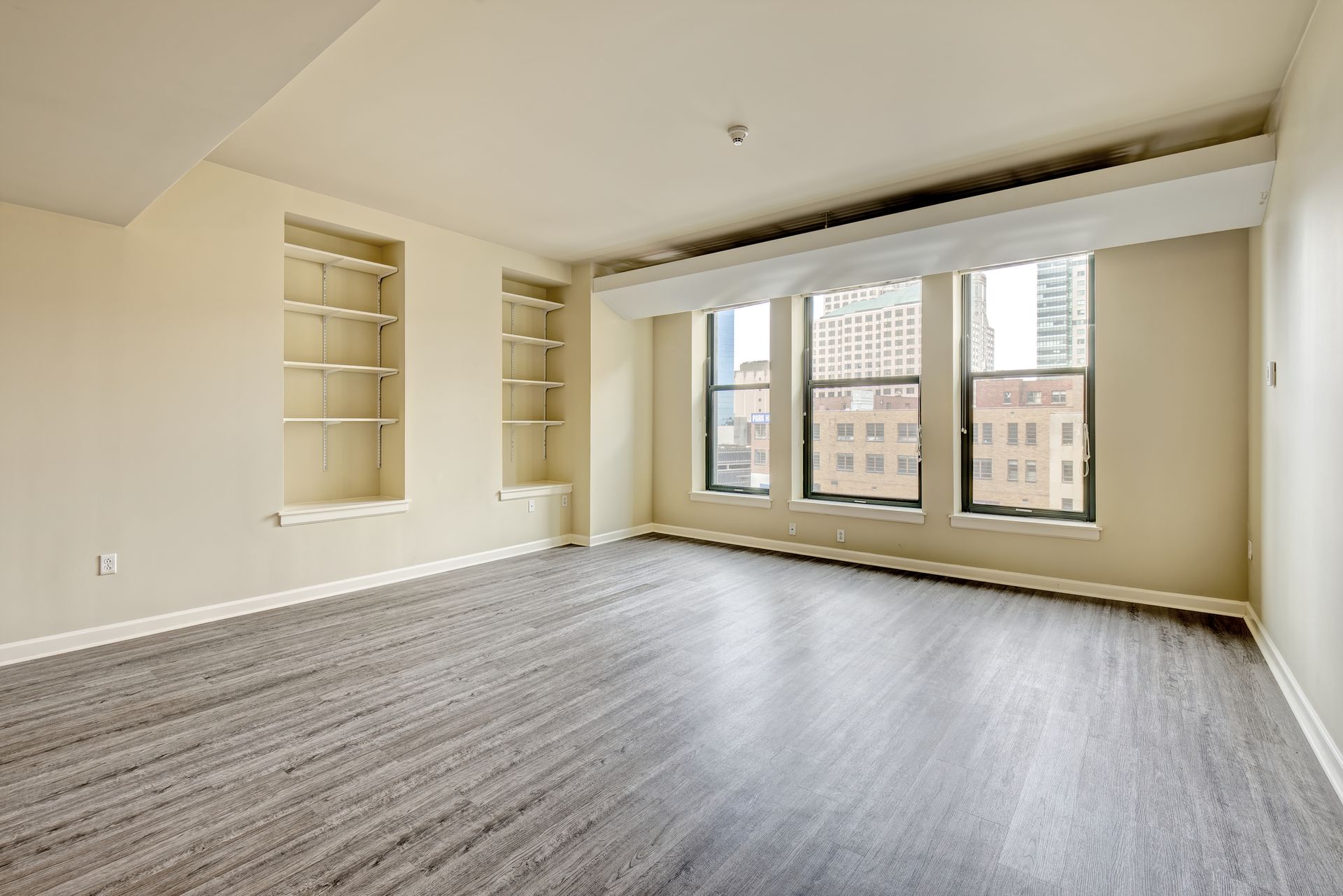 Empty living room with large windows overlooking city buildings, built-in shelving, and patterned flooring.