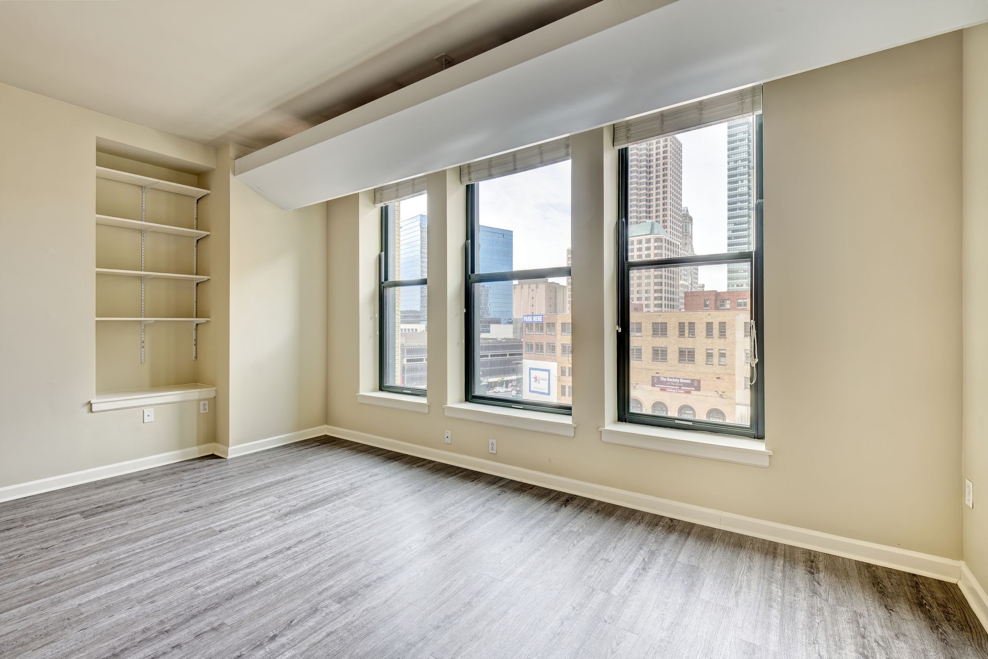 Empty room with large windows overlooking a city. The room has wood flooring, a built-in bookshelf, and neutral-colored walls.