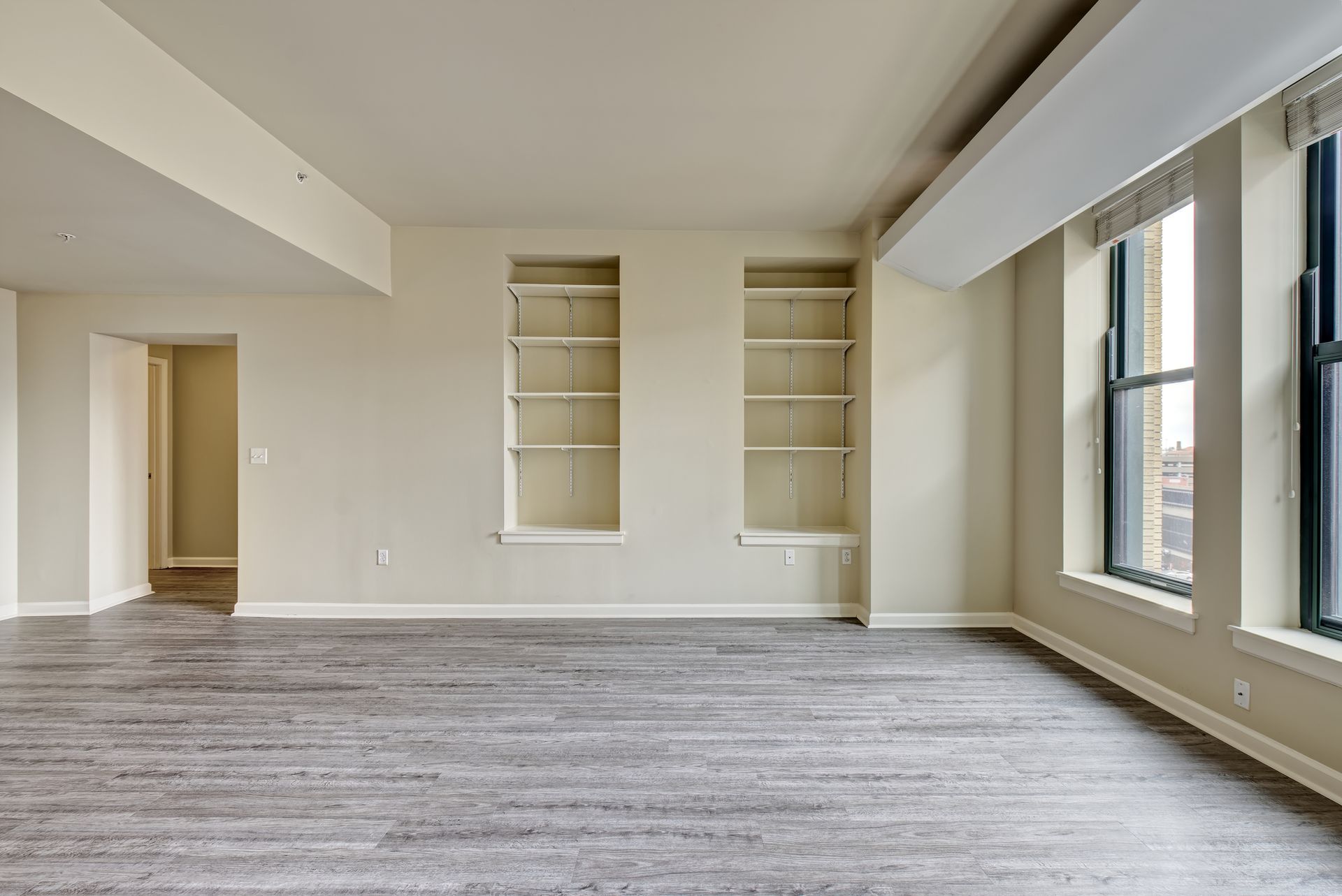 Empty room with grey wood-look flooring, built-in shelving, and large windows; beige walls and ceiling.