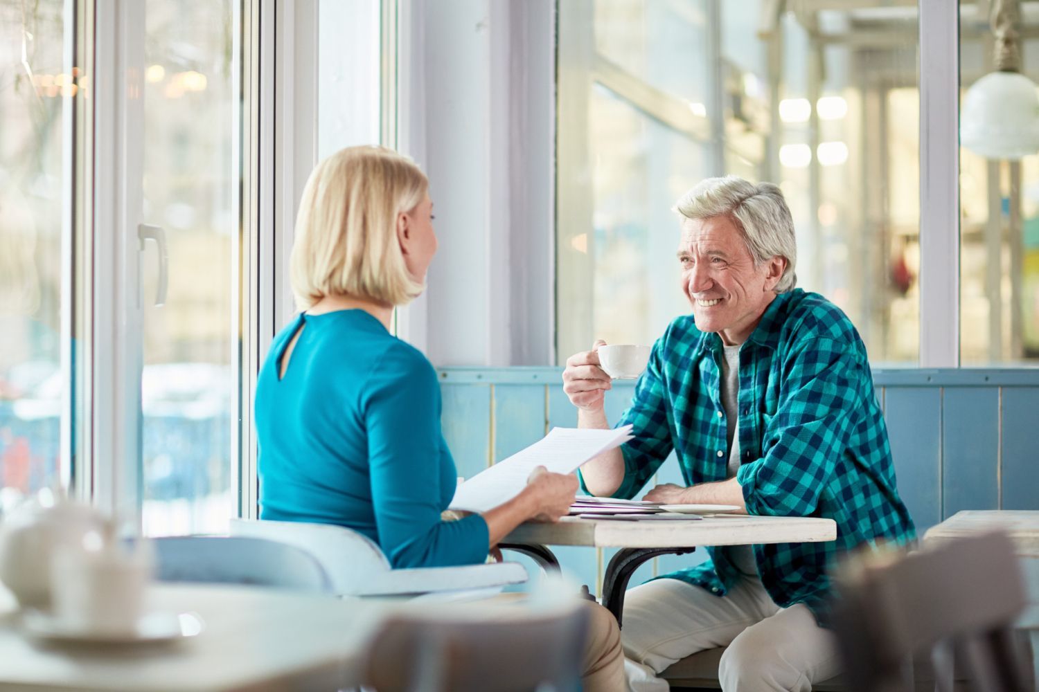 A woman in a teal shirt reviews papers with a smiling man in a plaid shirt, both at a table in a cafe.