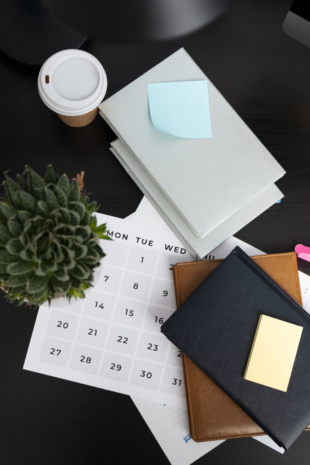 A desk scene with a stack of books, a coffee cup, a succulent plant, and a calendar. A sticky note is on the book.
