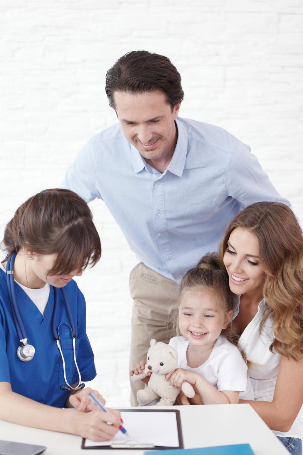 Doctor examining a smiling young girl with her parents; doctor is wearing blue scrubs and writing on a clipboard.