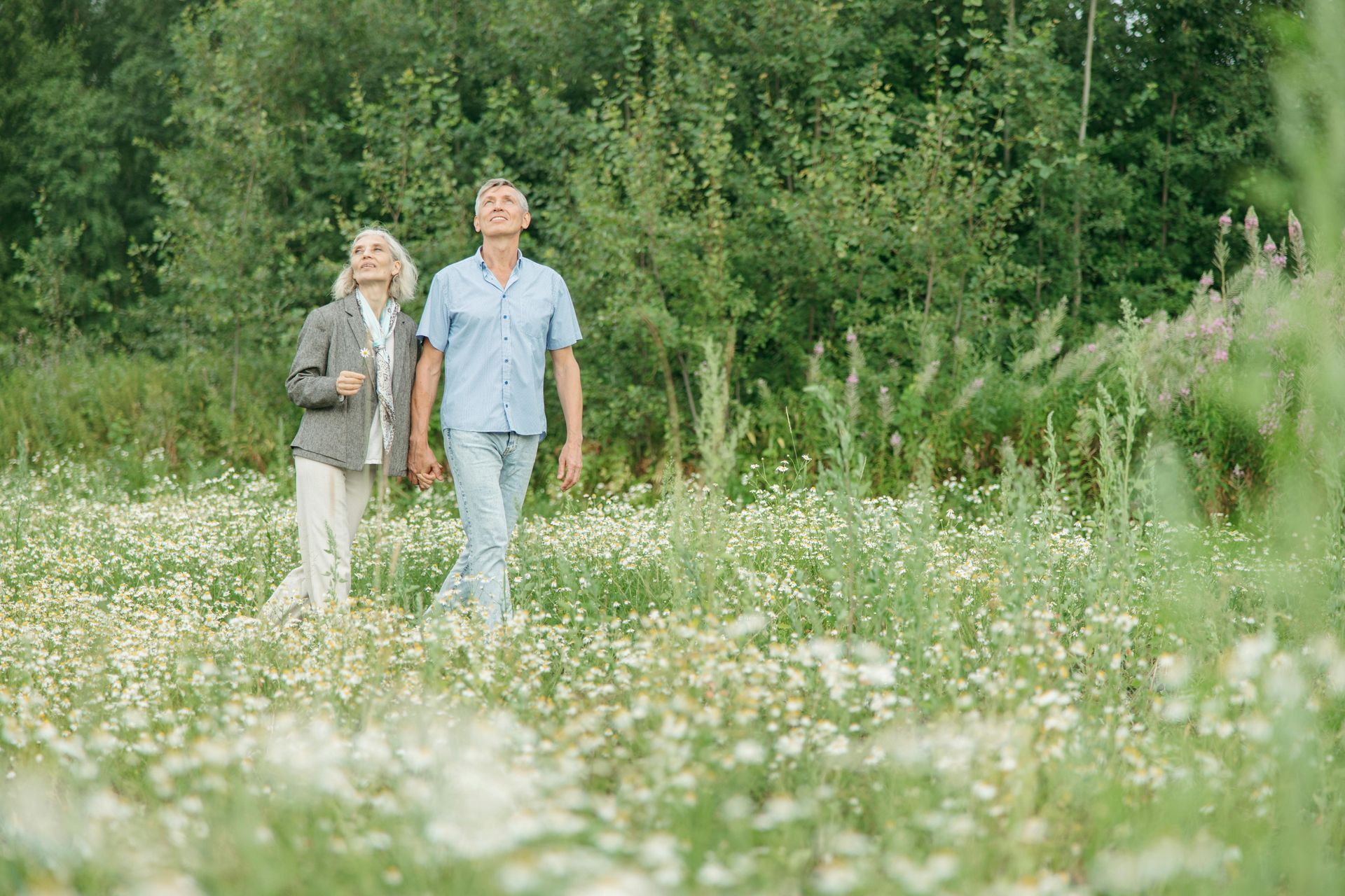 Two people walk in a field of white flowers, looking up. A forest is in the background.