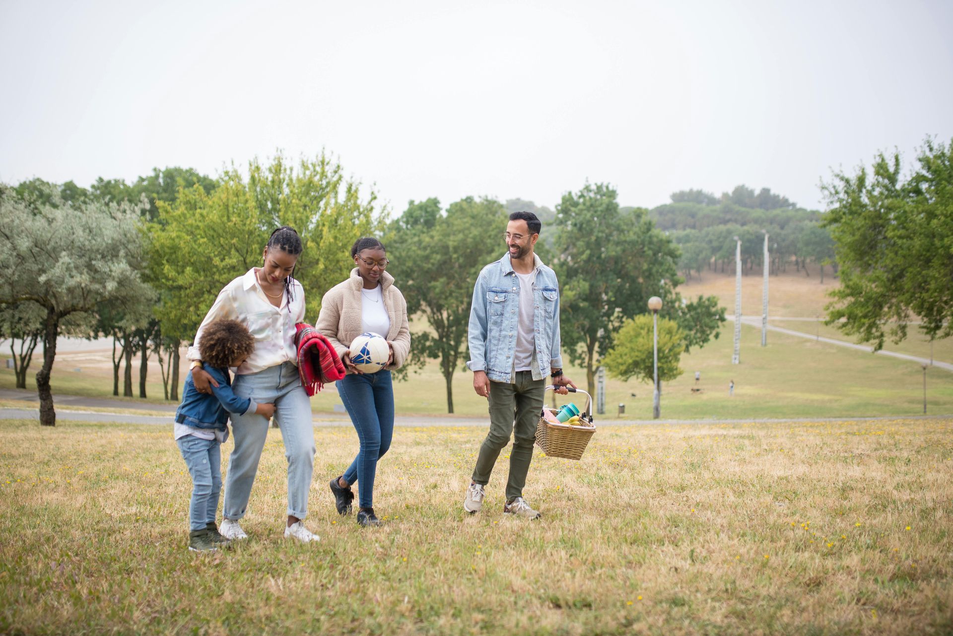 A family, including two parents and two children, walks through a grassy park, carrying a picnic basket and a ball.