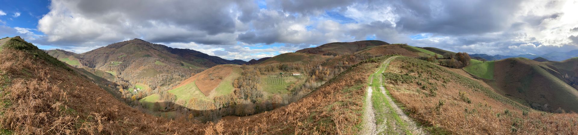 Panoramic view of rolling hills covered in brown vegetation under a cloudy sky. A path winds through the center.