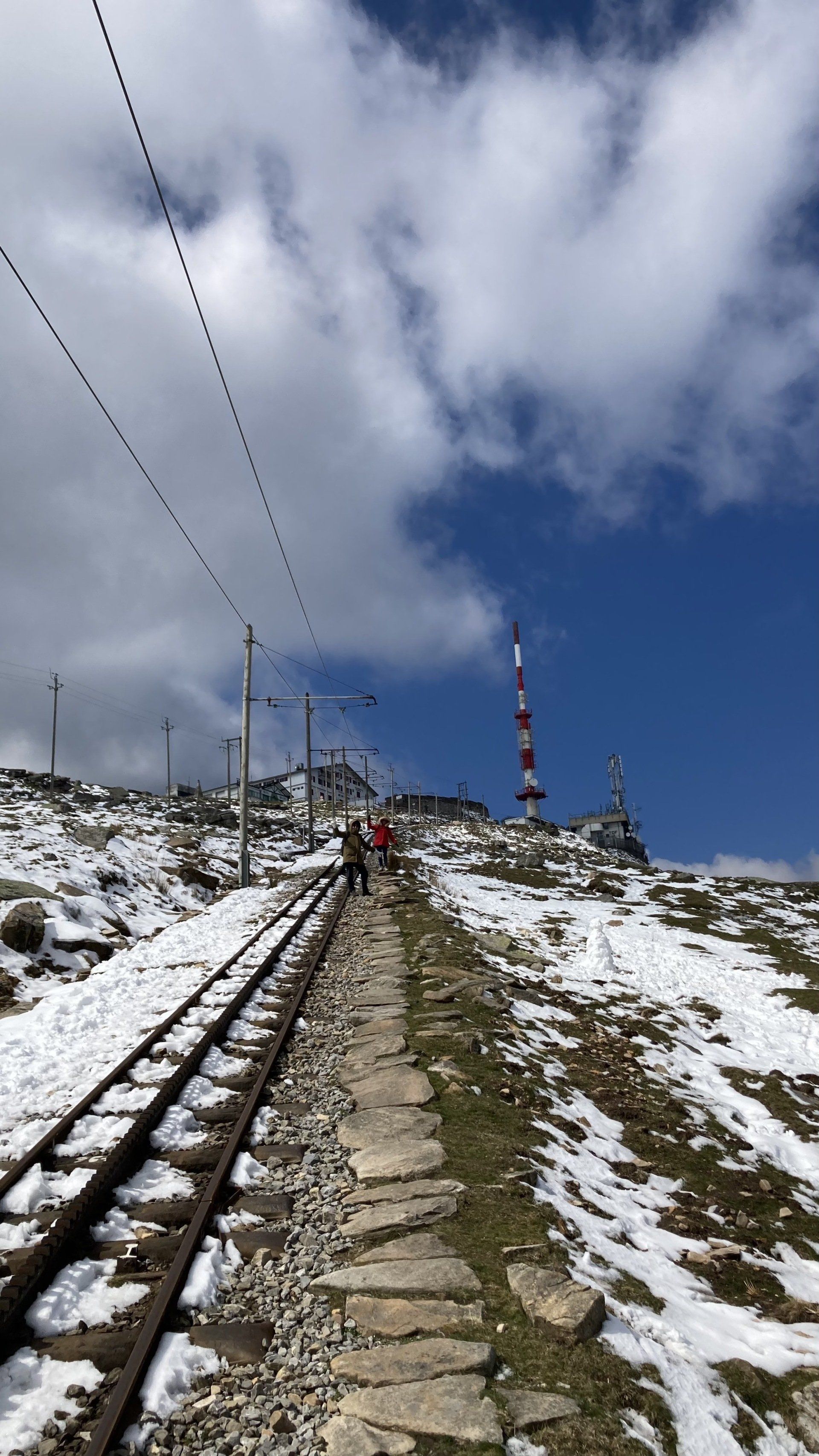 Snowy mountain path with train tracks leading to buildings under a blue sky with clouds.