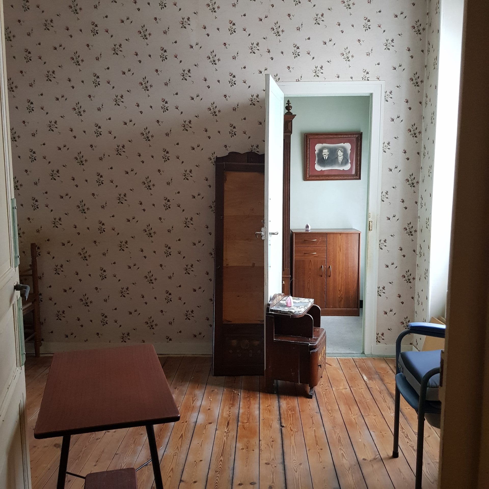 sitting room before renovation with old fashioned wallpaper, scattered pieces of old furniture, exposed wooden floors