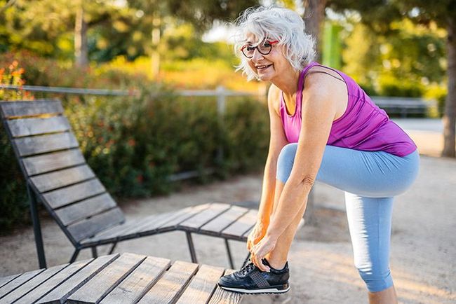 Woman stretching beside a park bench, bending to touch her foot in a sunny outdoor setting