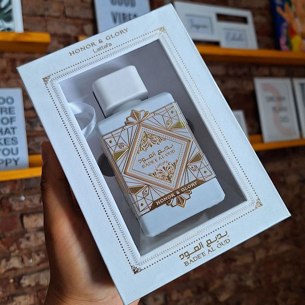 Mano sosteniendo una caja de regalo de perfume blanca con diseño geométrico dorado y frasco, sobre un fondo de ladrillos.