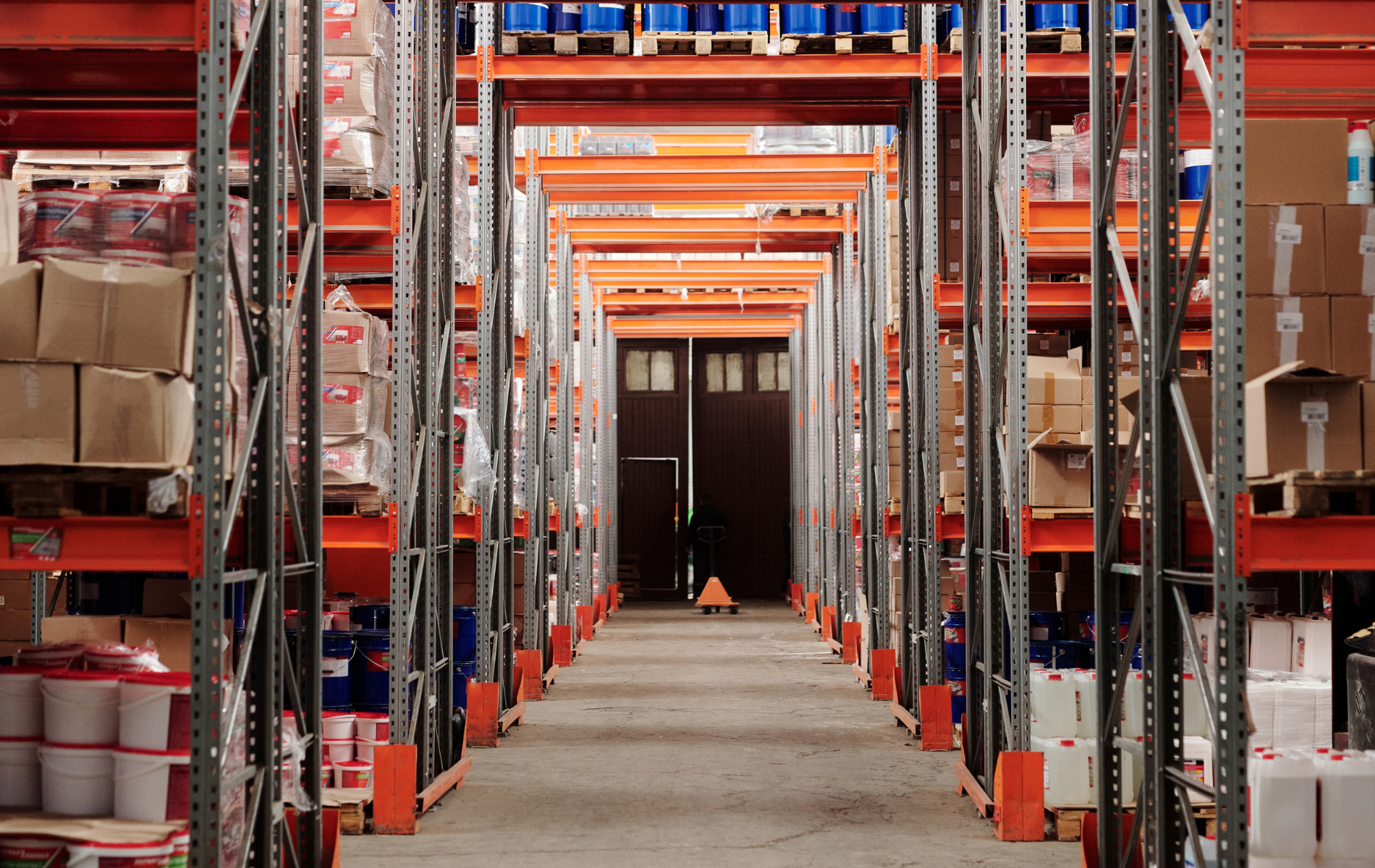 Warehouse aisle lined with shelves of boxes and supplies, viewed from the center toward a distant doorway