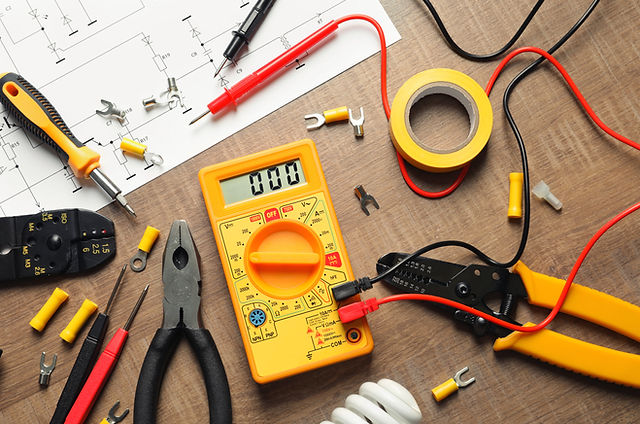 Yellow multimeter on a workbench with pliers, wires, tape, and electronics tools