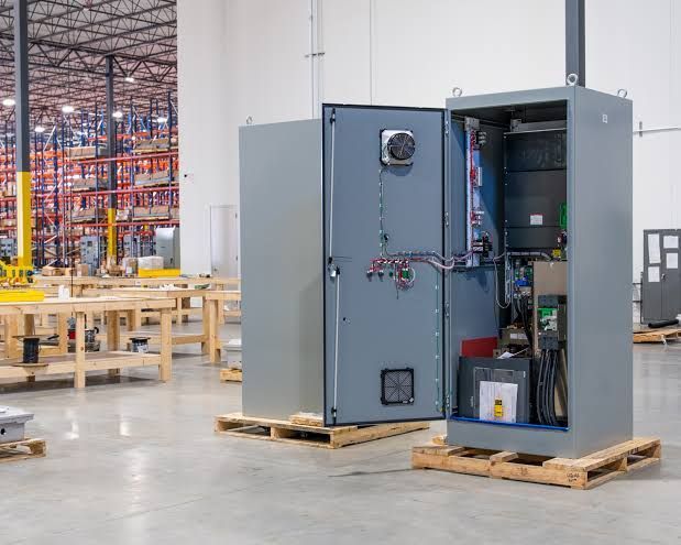 Open electrical cabinet on pallets in a warehouse, with shelves and industrial equipment in the background