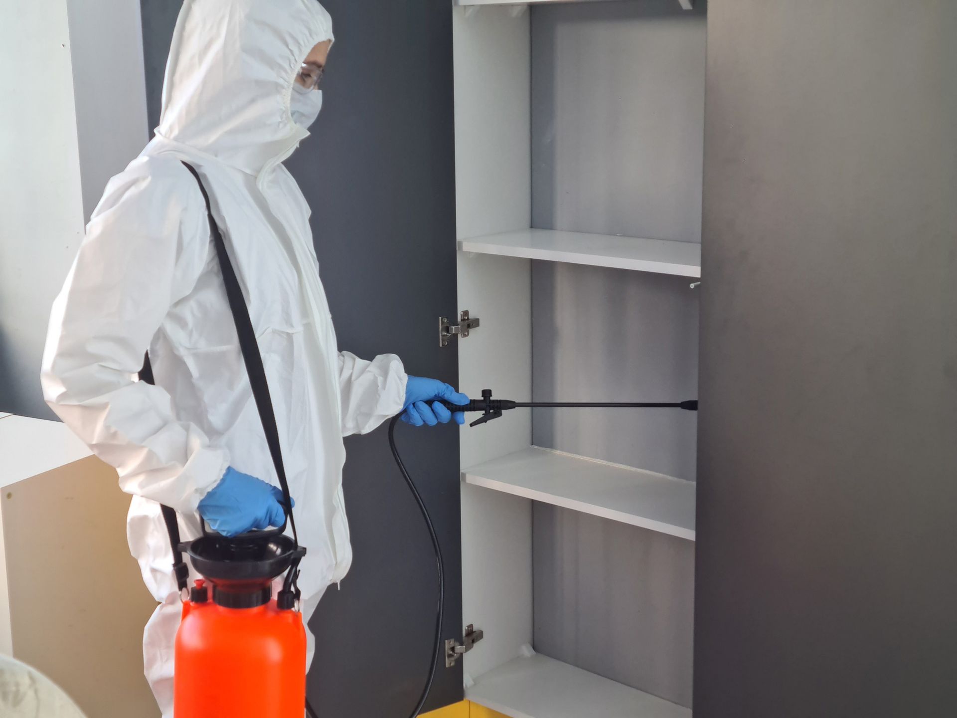 Technician in protective gear sprays inside cabinet during pest control treatment. Technician in protective gear sprays inside cabinet during pest control treatment.