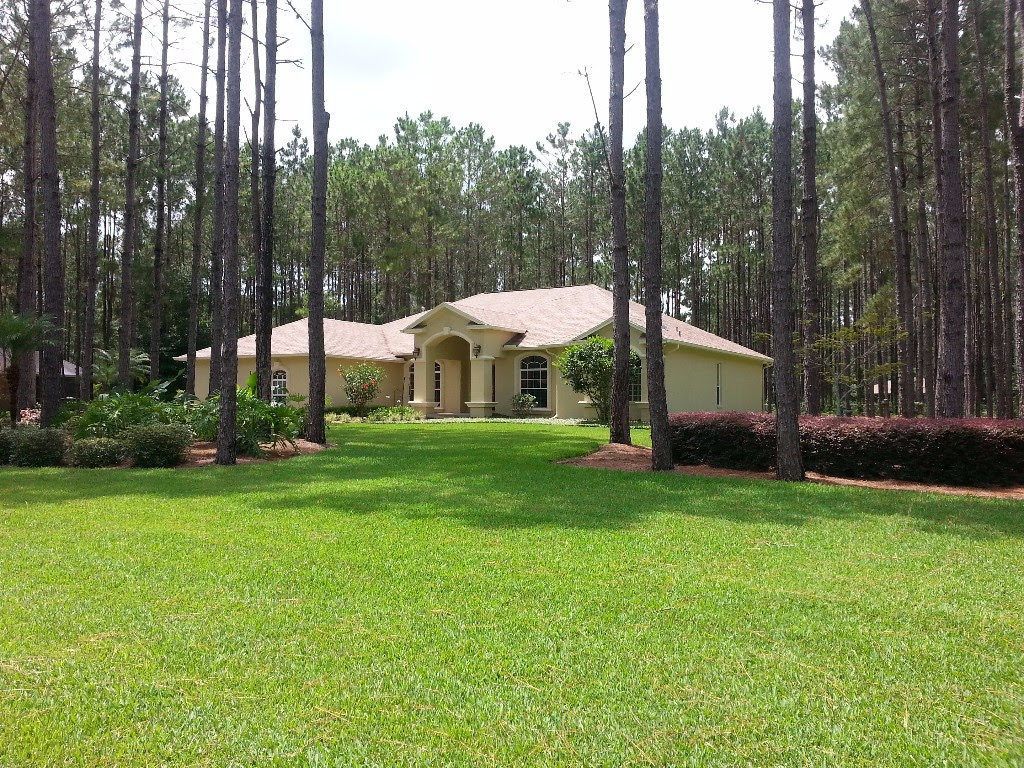 House with beige siding, brown roof, surrounded by tall pine trees and green lawn.