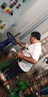 Dos personas en un patio industrial; una de ellas, con mono de trabajo, señala una válvula de tubería mientras la comenta con un compañero.