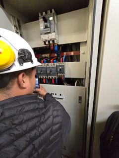 Un trabajador con casco y orejeras realiza el mantenimiento de un panel de control rojo de la alarma contra incendios, situado junto a un anuncio de juegos de azar.